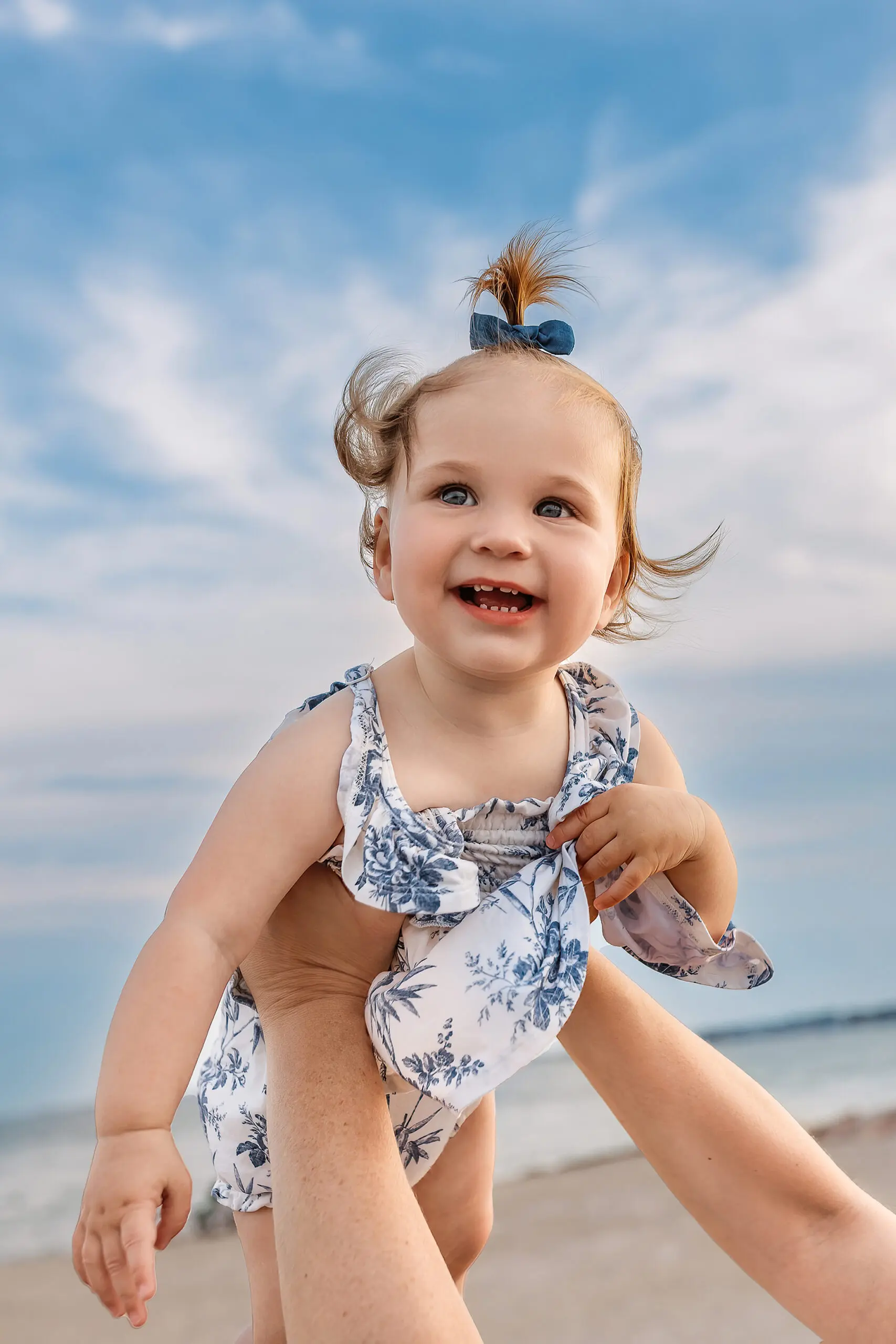 A smiling toddler with light brown hair and a blue bow is held up in the air by an adult’s hands at the beach. She wears a white and blue floral outfit, with a blue sky and clouds in the background.