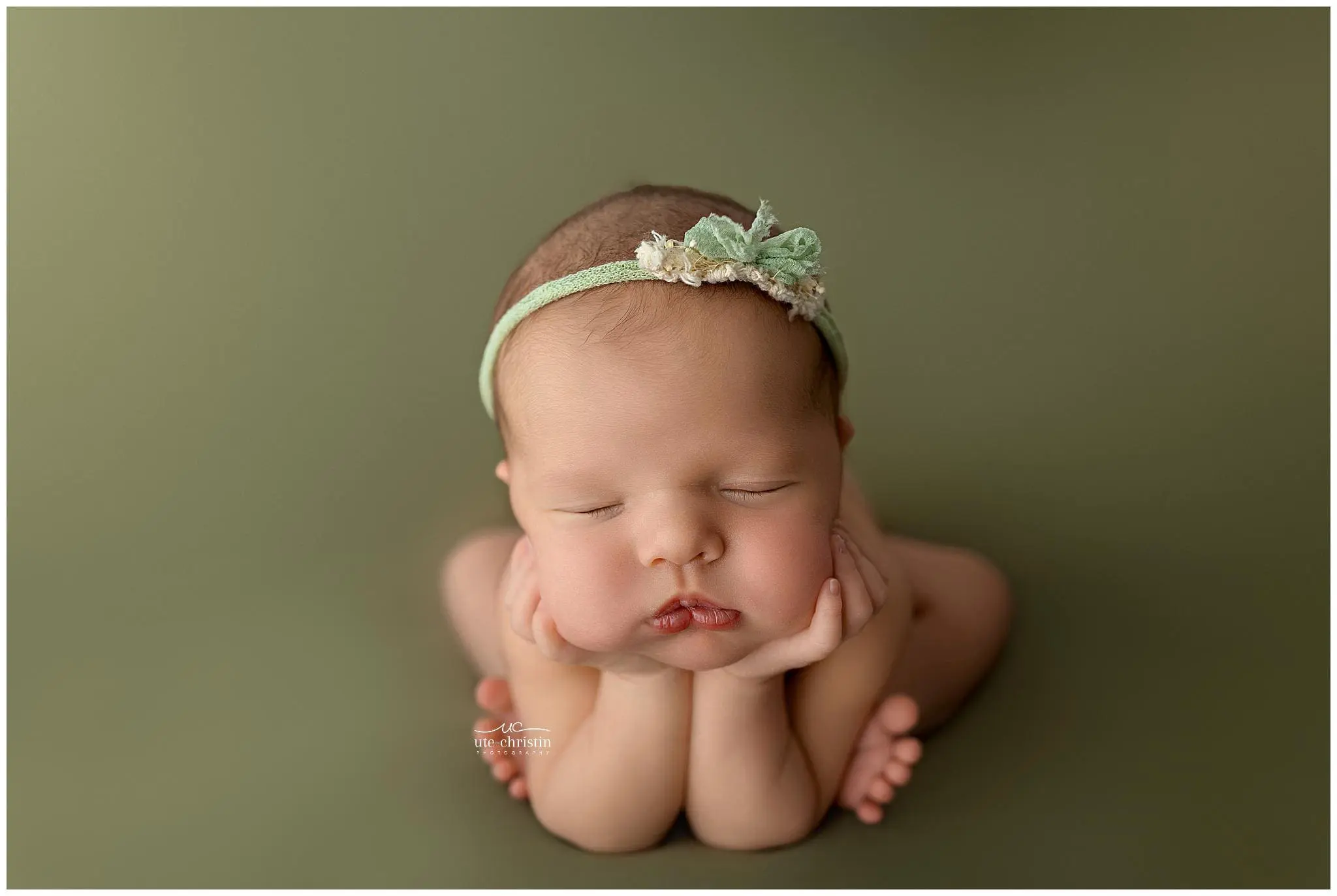 A newborn session in New Haven County features a baby with eyes closed, chin resting on hands, elbows propped up, wearing a green floral headband and posed against a soft olive green background.