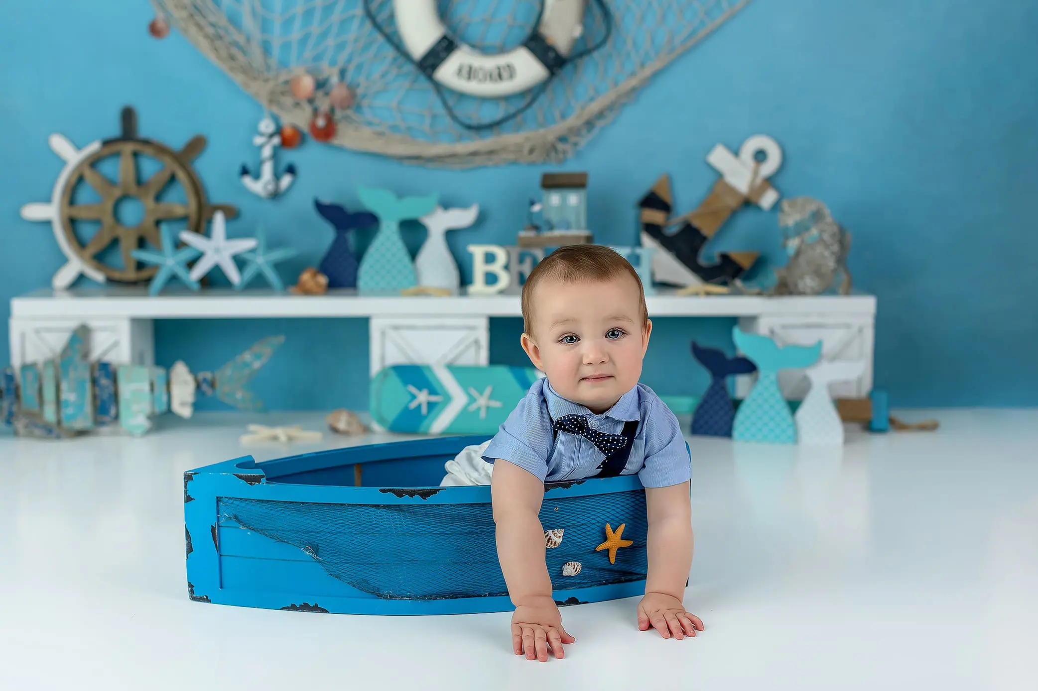 A baby in a blue shirt with a bow tie sits charmingly in a small wooden boat. Surrounding him are nautical decorations like steering wheels, anchors, and fish designs, all in serene blue and white tones during his Connecticut cake smash..