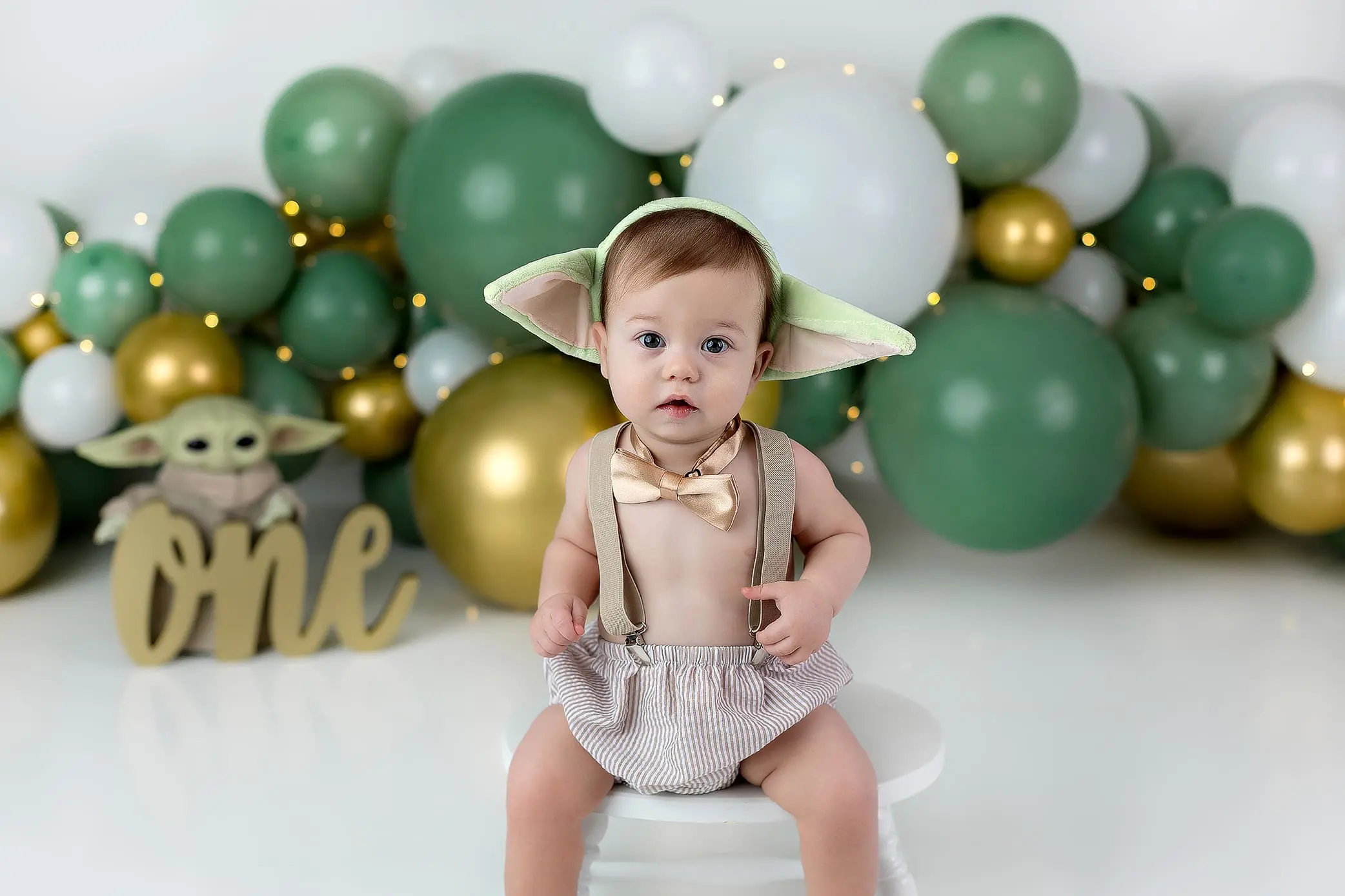 A baby wearing a Yoda-inspired hat and suspenders sits on a stool. The background features green, white, and gold balloons with a gold one sign and a Yoda plush toy. The baby is dressed for a first birthday celebration in CT.