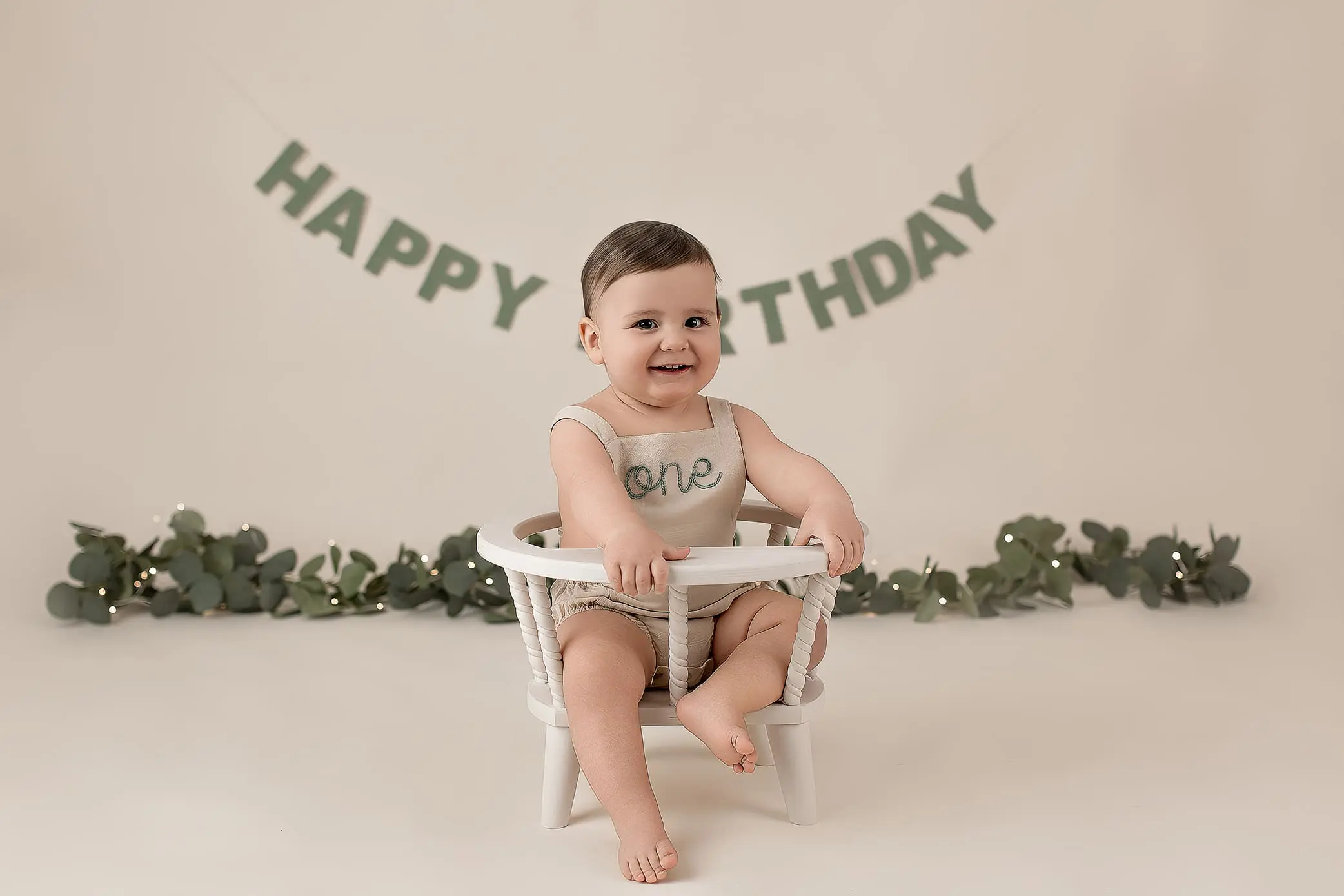 A smiling baby sits in a white chair, wearing a light-colored outfit with one on it. In the background, a Happy Birthday banner hangs above greenery. The setting is soft and neutral-toned, celebrating the babys first birthday.