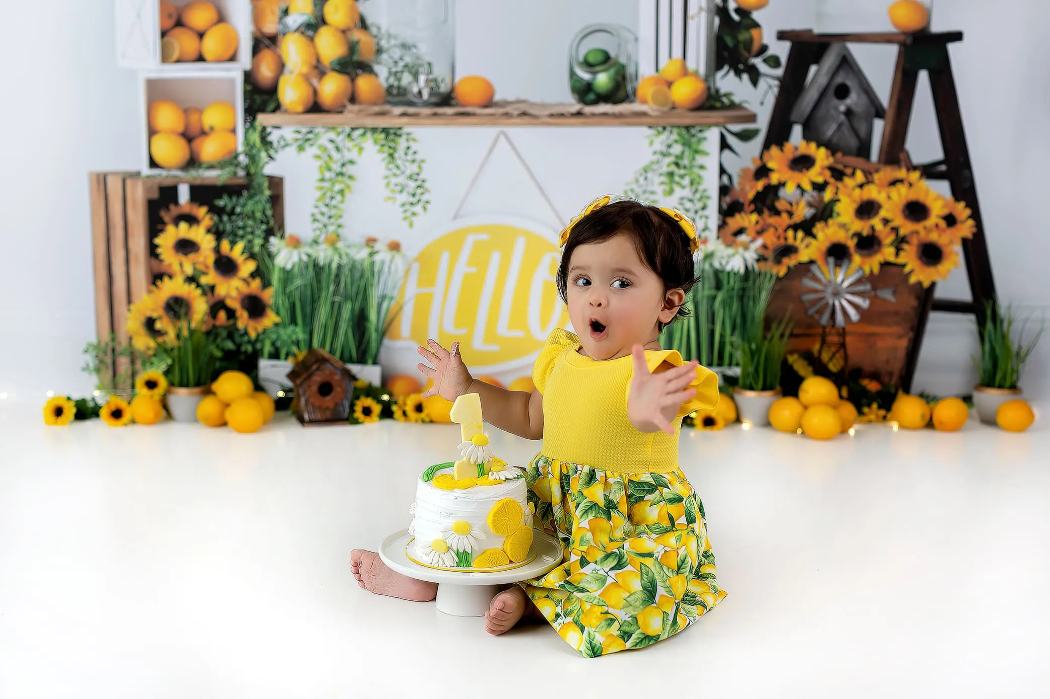 A toddler in a yellow top and floral skirt sits on the floor, surprised and joyful during her cake smash. In front of her is a cake adorned with lemon decorations. The background features a cheerful lemon and sunflower theme.