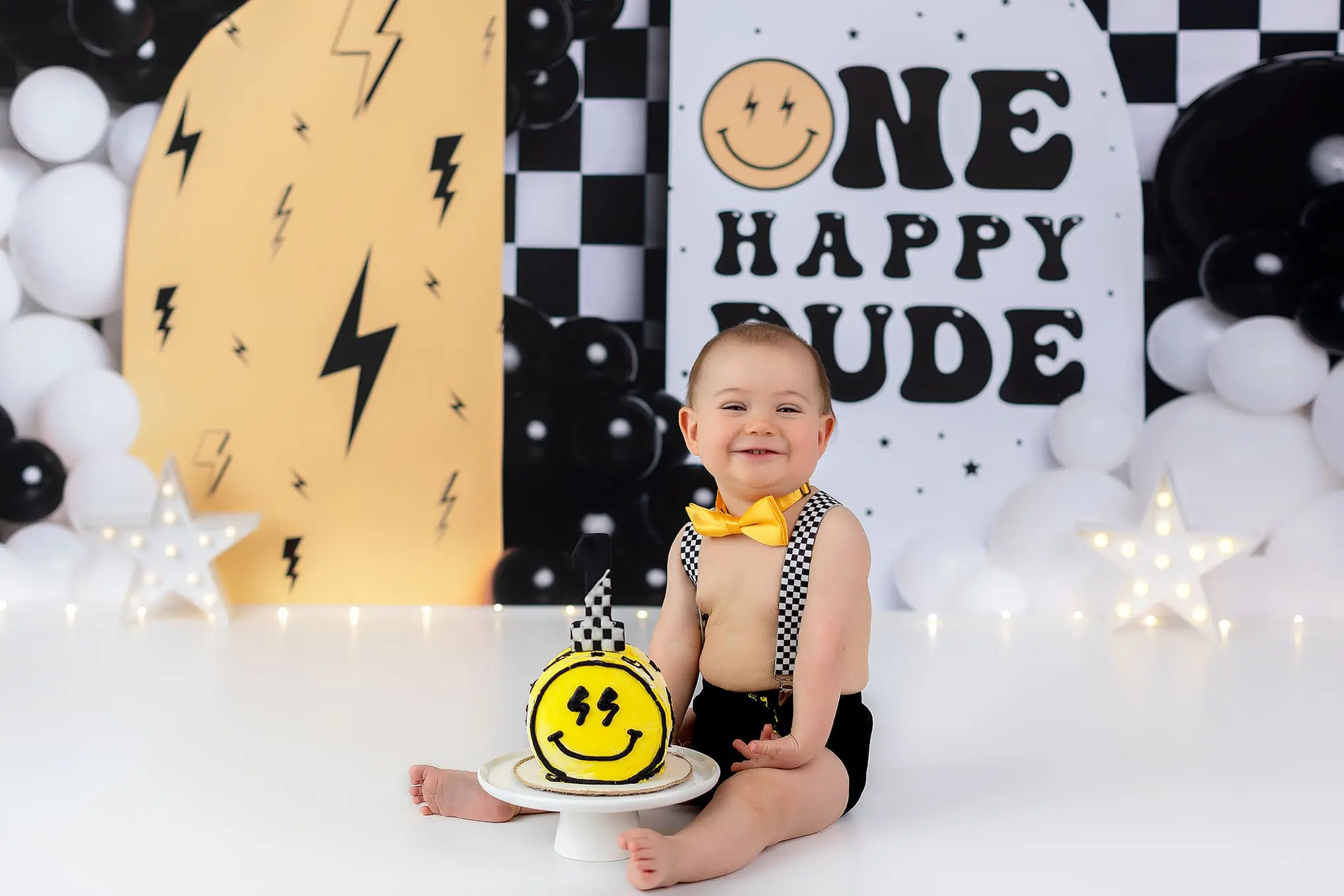 A baby sits on the floor, smiling, wearing a yellow bow tie and checkered suspenders. In front is a yellow cake with a smiley face. The background features black, white, and yellow balloons, with signs reading One Happy Dude and a star decoration.