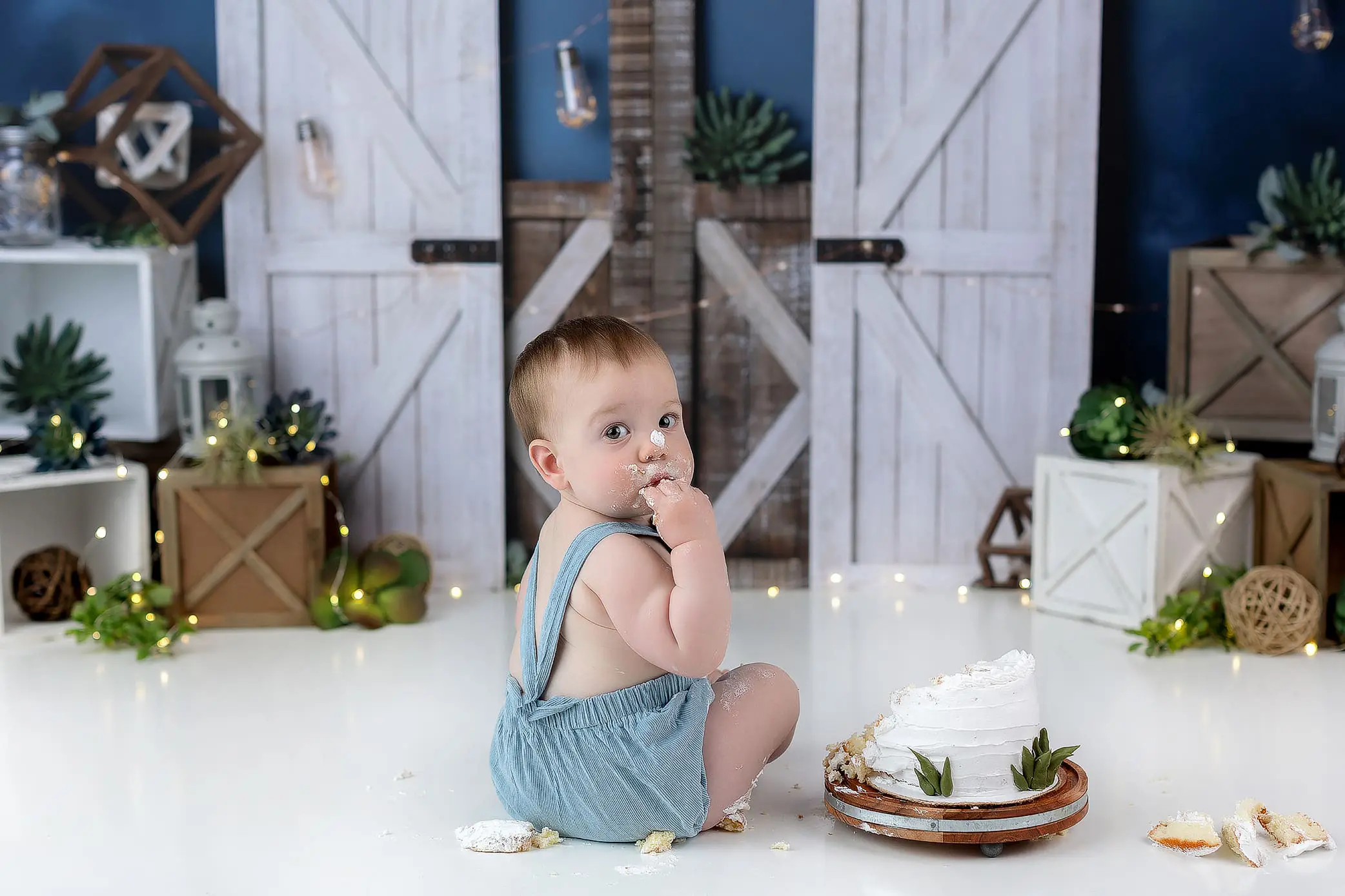 A baby in blue overalls sits on the floor, facing backward, delighting in cake with messy hands. Before them lies a partially eaten white cake. Rustic decor with plants and fairy lights frames this charming moment of Connecticut kids boundless joy.