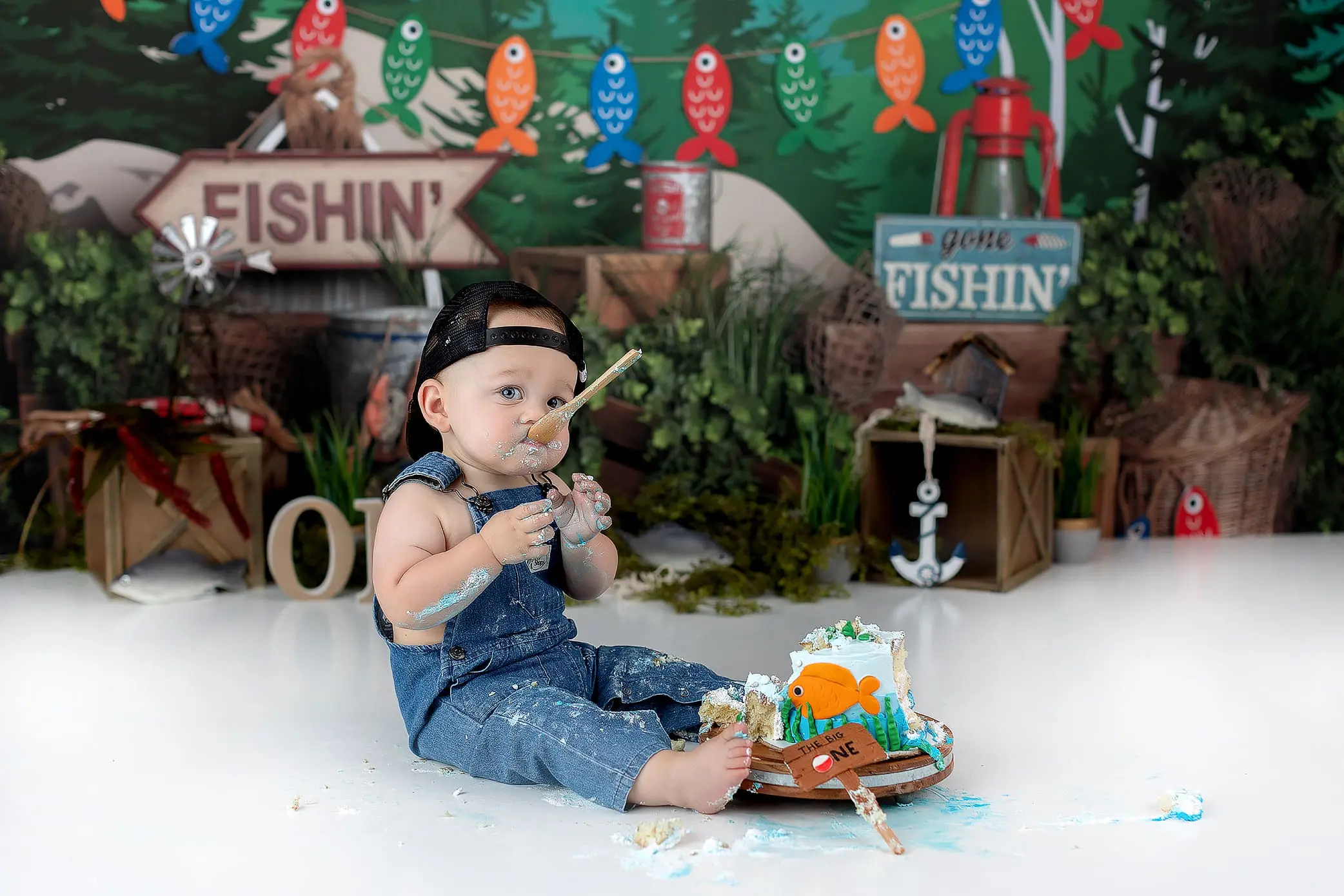 A baby in a blue denim outfit and cap sits on the floor, surrounded by a forest and fishing-themed backdrop. The baby is eating cake, with frosting on their face and hands. Nearby props include a Gone Fishin sign, fishing baskets, and colorful fish decor.