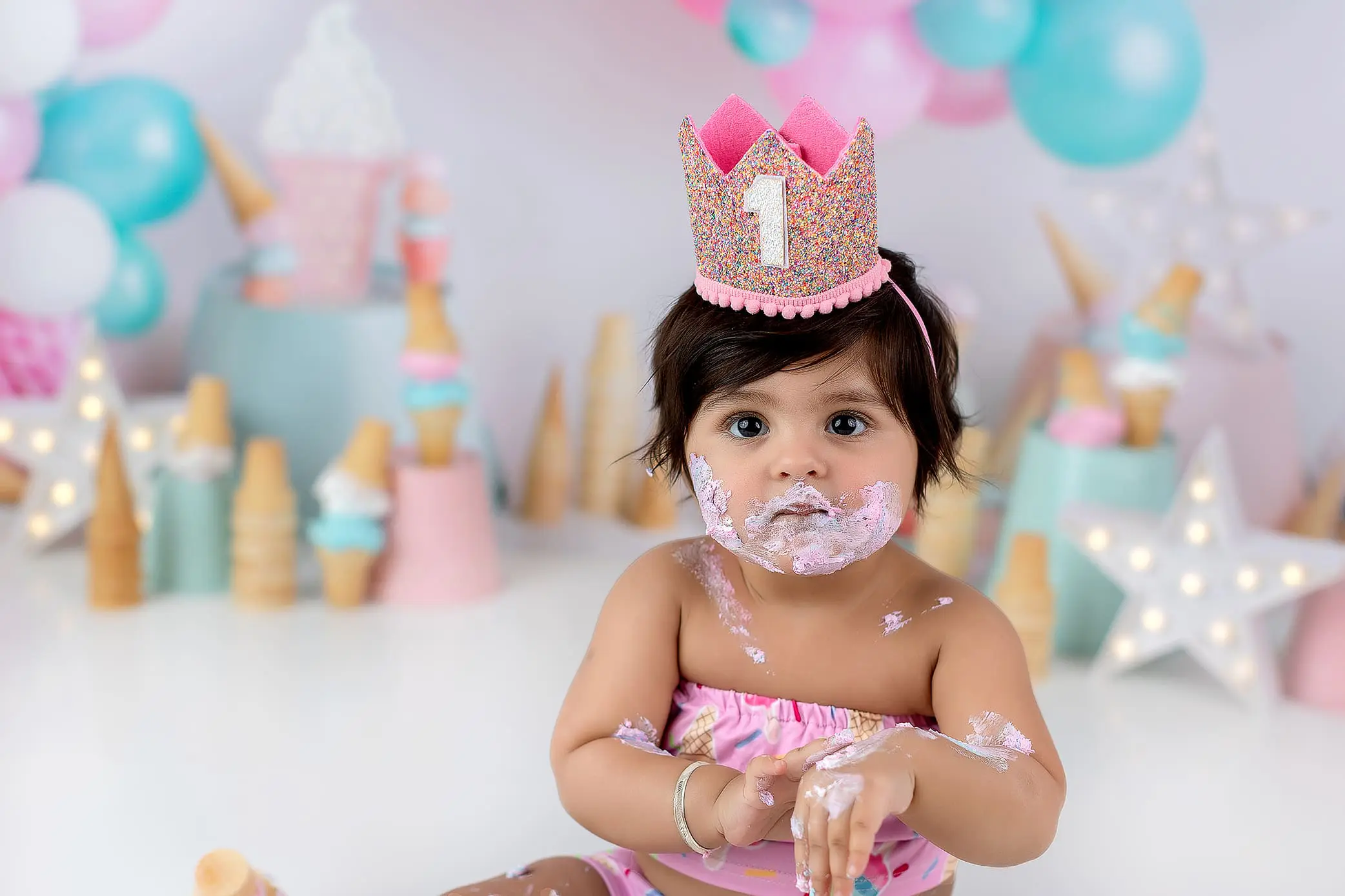 A baby with dark hair and a crown labeled 1 sits surrounded by pastel balloons and ice cream-themed decorations. The baby is covered in cake and frosting, wearing a pink outfit, with a curious expression in a festive cake smash setting.