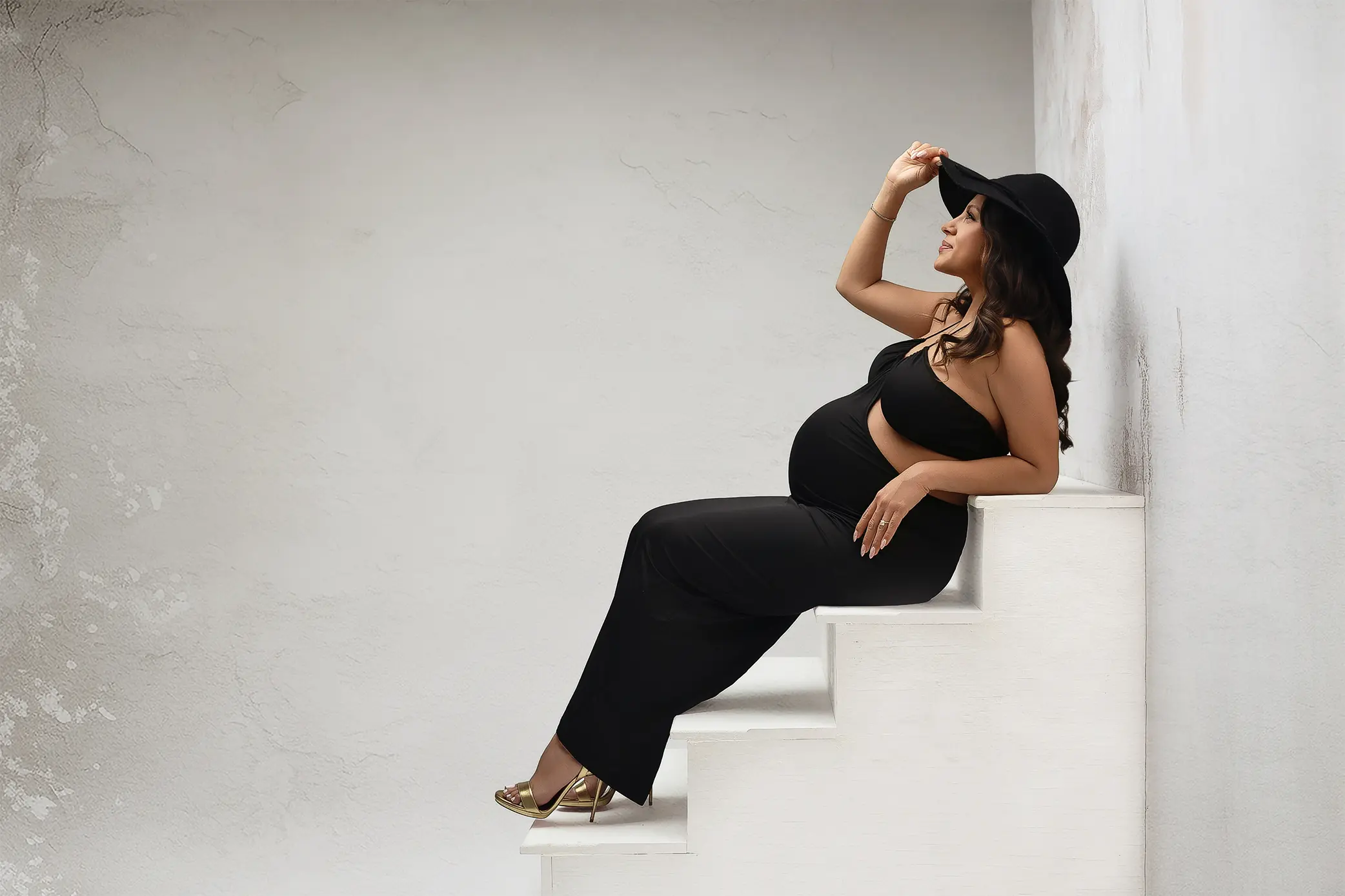 A pregnant woman in a fitted black dress, gold heels, and a wide-brimmed hat sits on white stairs, leaning back and touching her hat with a serene expression against a plain light background during her CT maternity session.