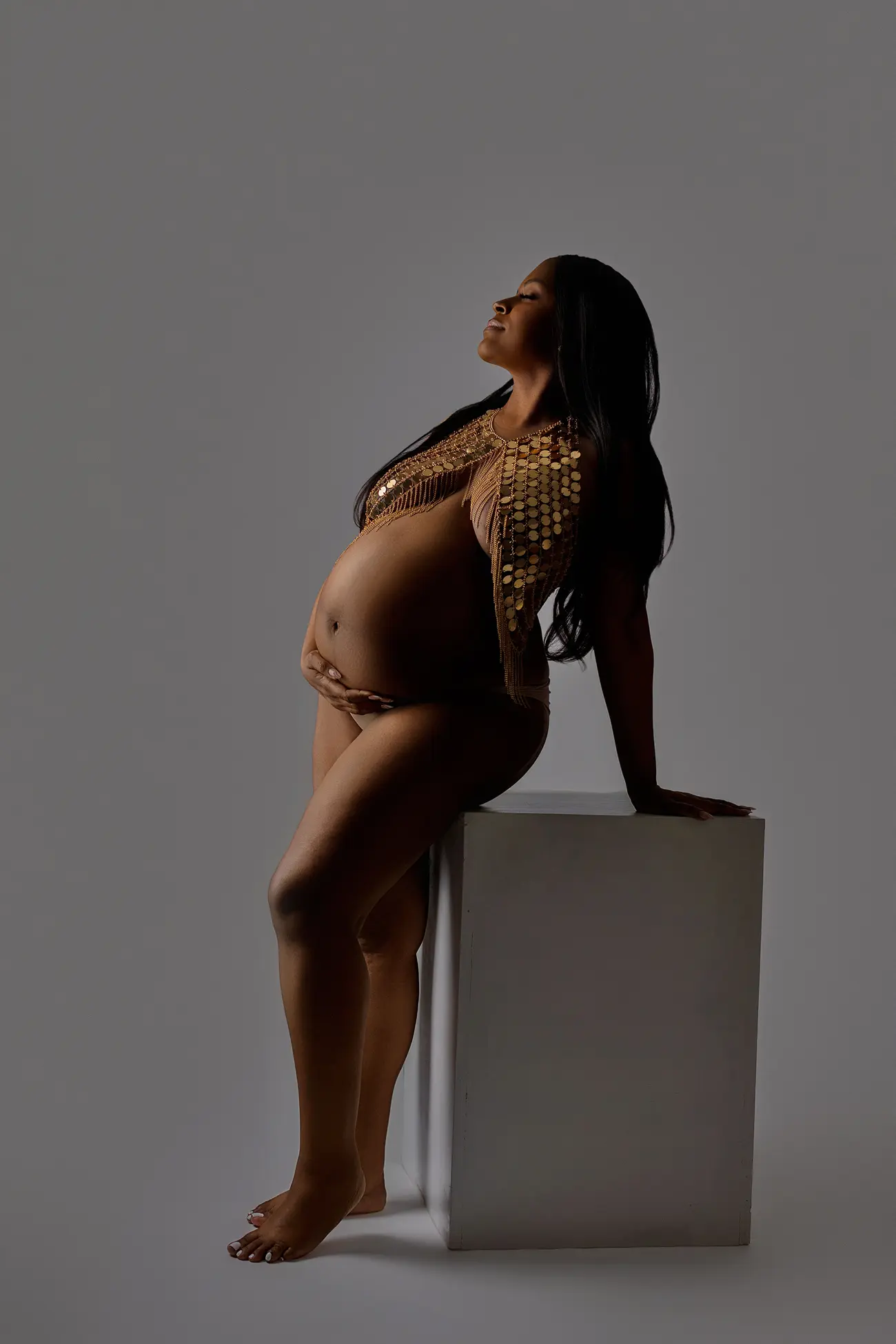 A pregnant woman poses gracefully, sitting on a white block and looking upward. She wears a gold embellished top and cradles her belly, with a neutral gray background.