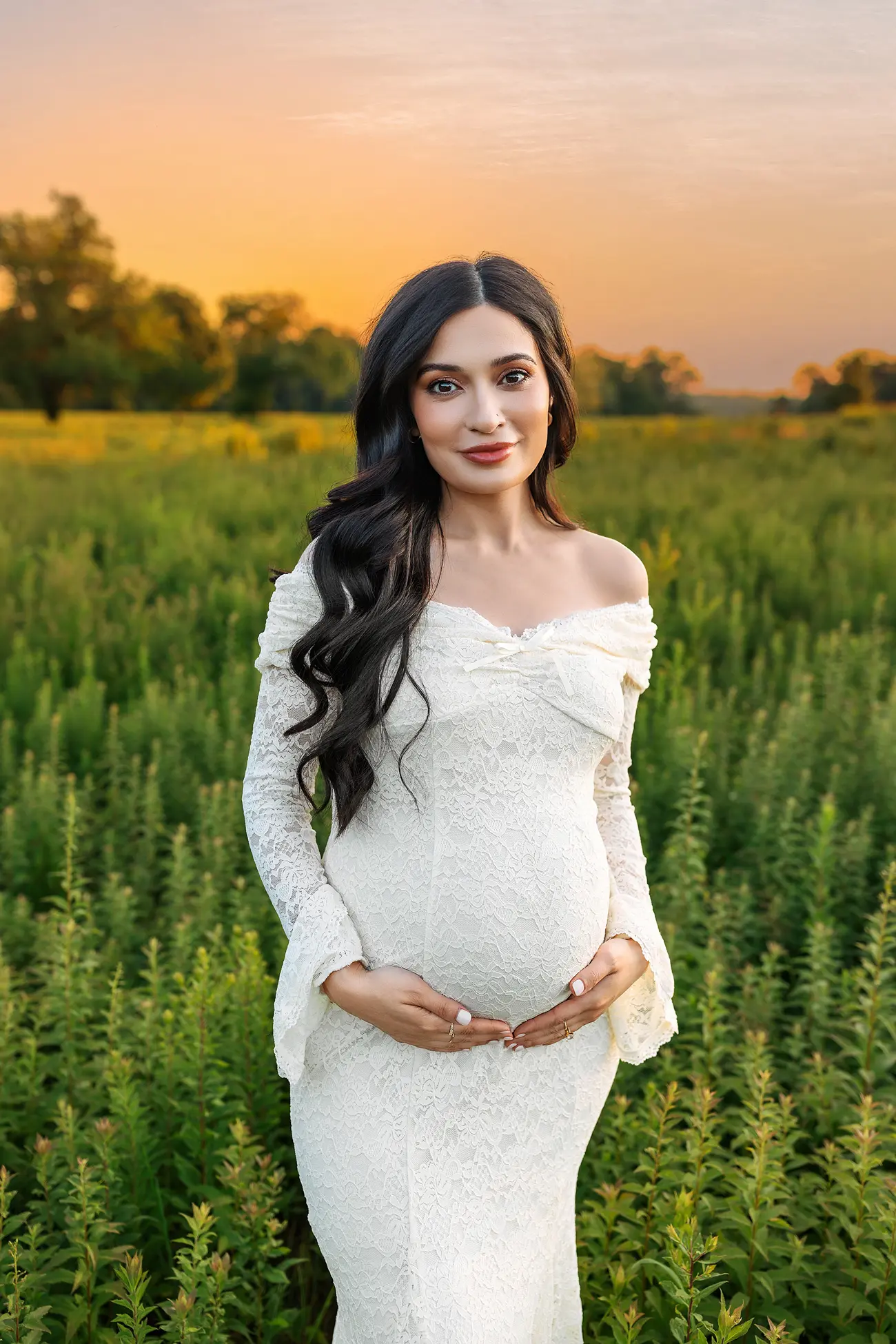 A pregnant woman in a white lace dress stands in a green field at sunset, gently holding her belly and smiling at the camera.