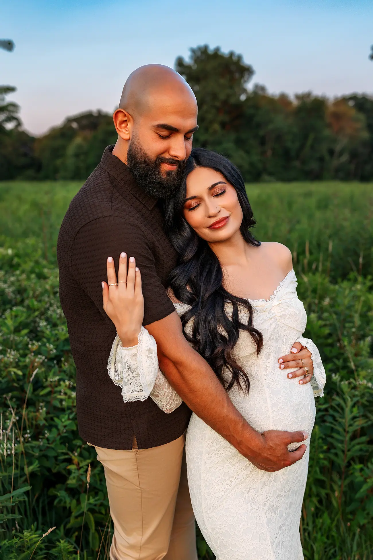 A couple stands in a green field. The man hugs the woman from behind, his hands on her pregnant belly. The woman wears a white dress and rests her head on his chest, both look peaceful and content.