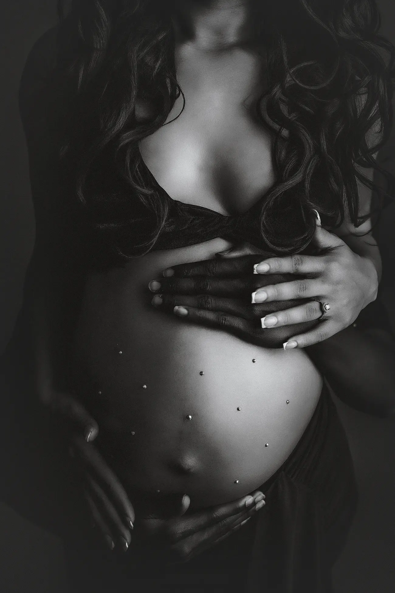 A black and white photo of a pregnant woman holding her bare belly, adorned with small jewels. She wears a dark bra and drapes, her long hair falling over her shoulders. One hand displays a ring.