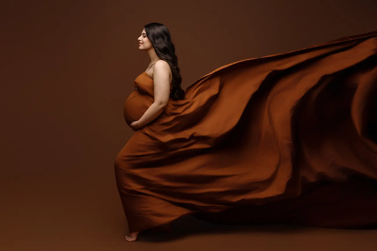 A pregnant woman in a flowing, rust-colored gown stands barefoot against a brown background. Perfect for maternity photos Milford, the dress billows dramatically as she cradles her belly and looks serene.