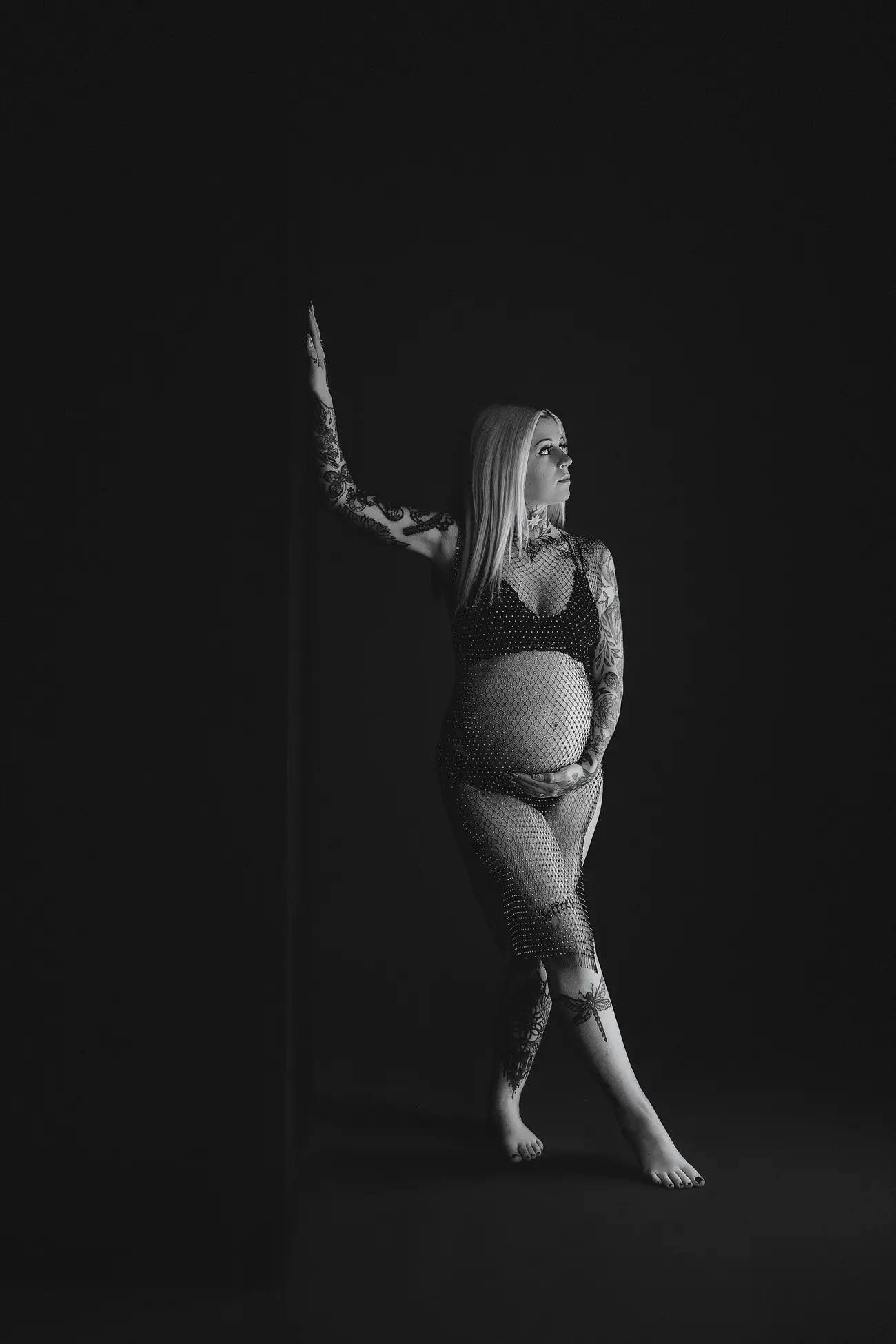 Black-and-white photo of a pregnant woman with tattoos, wearing a mesh outfit, standing barefoot and leaning against a dark wall. She looks to the side and rests one hand on her belly.