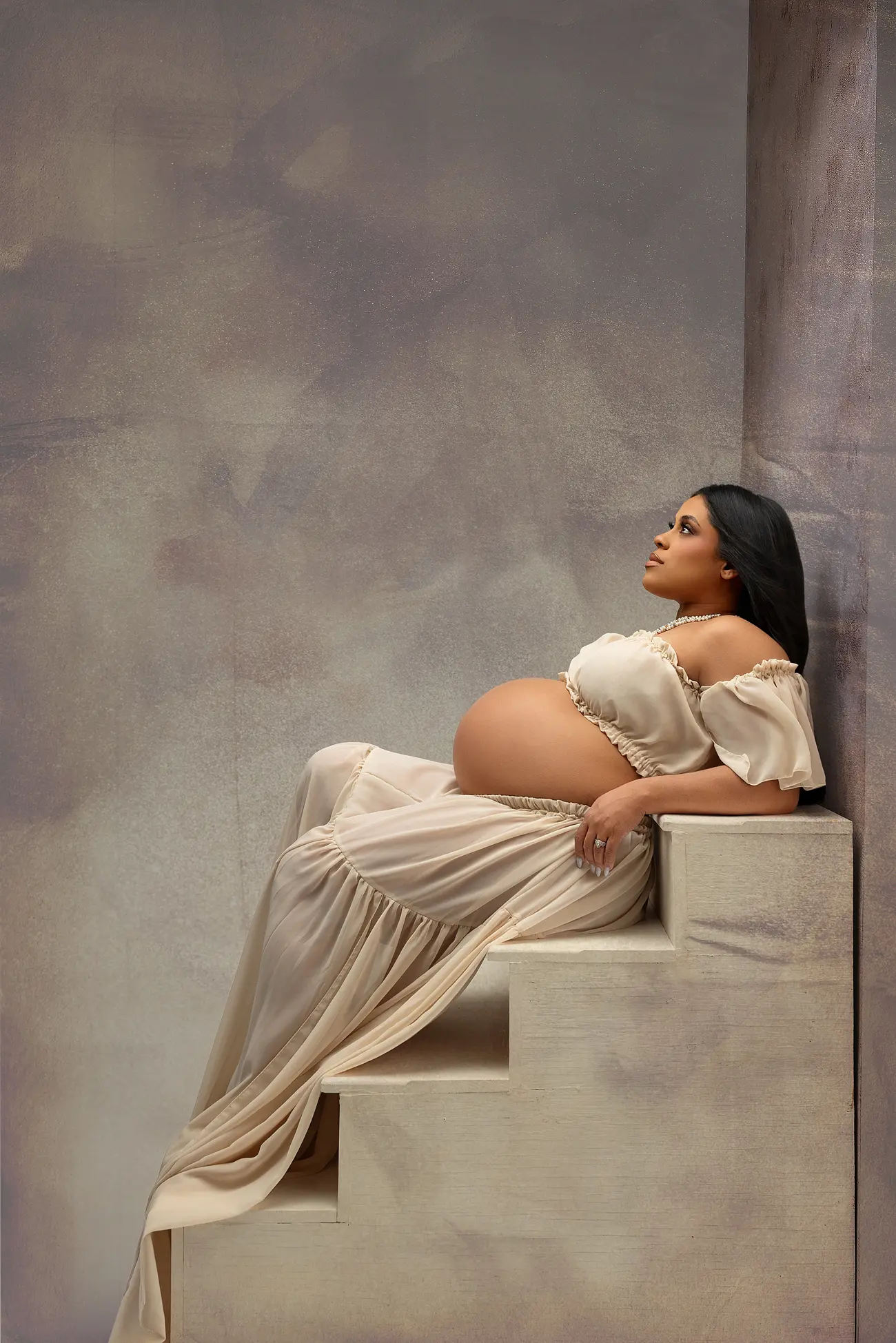 A pregnant woman in a flowing, sheer beige dress sits on steps, leaning back against a wall and gazing upward thoughtfully, captured by a CT maternity photographer amid soft lighting and a neutral, artistic background.