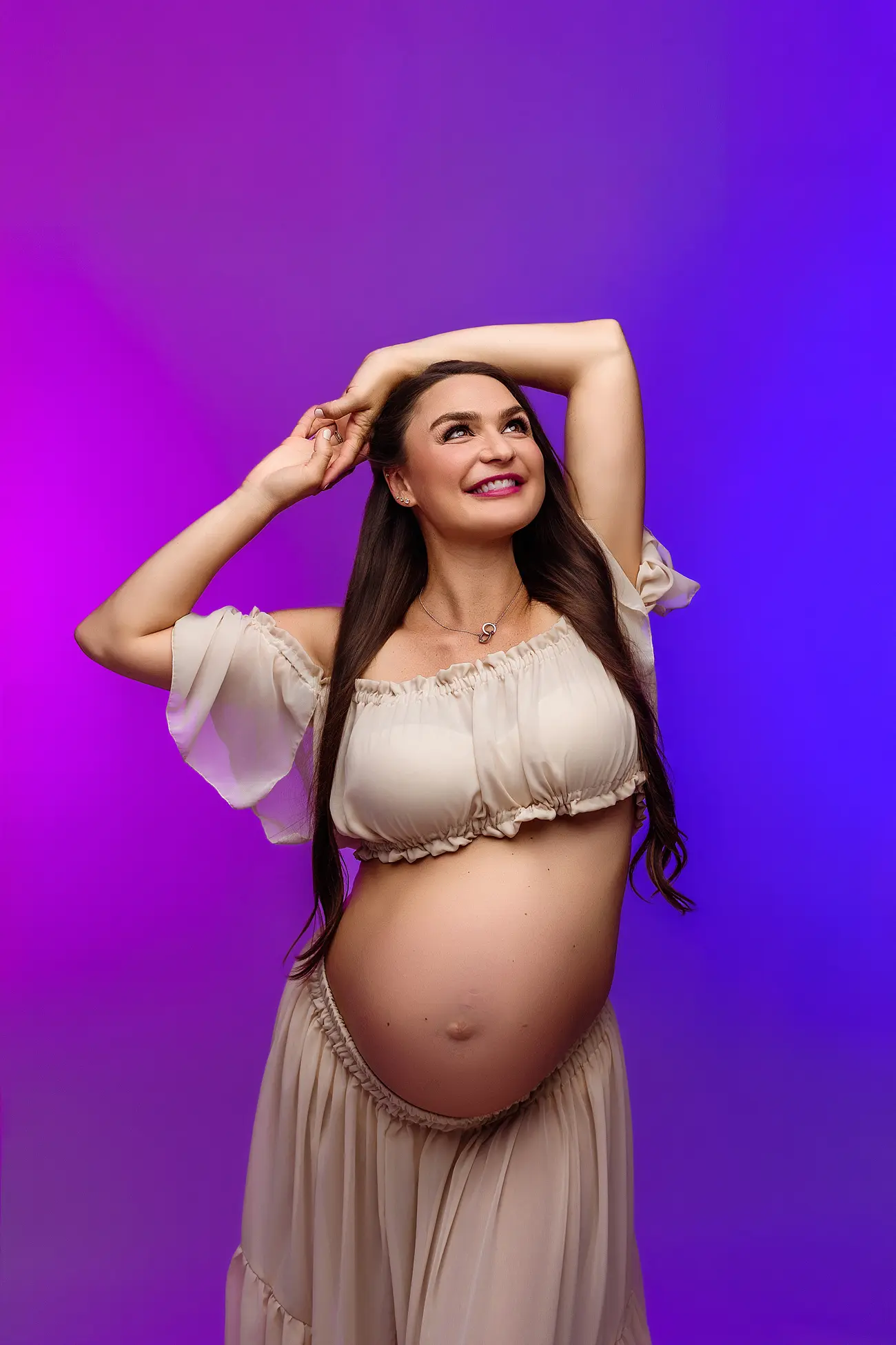 A pregnant woman in a flowing, off-shoulder beige dress poses smiling with her hands above her head against a vibrant purple and blue gradient background during a CT maternity shoot.