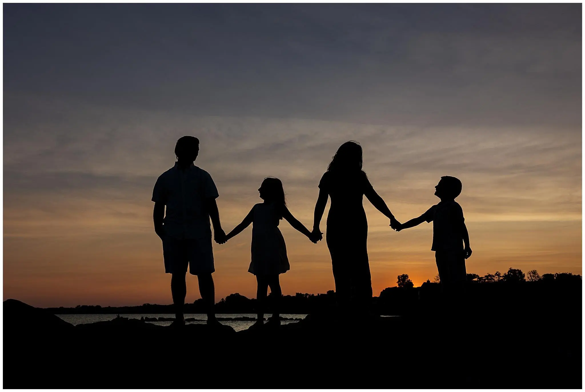 Silhouetted image of four people holding hands at sunset, standing against a colorful sky. Two adults are on the left, with two children on the right, forming a continuous line with their joined hands. The scene exudes unity and warmth.