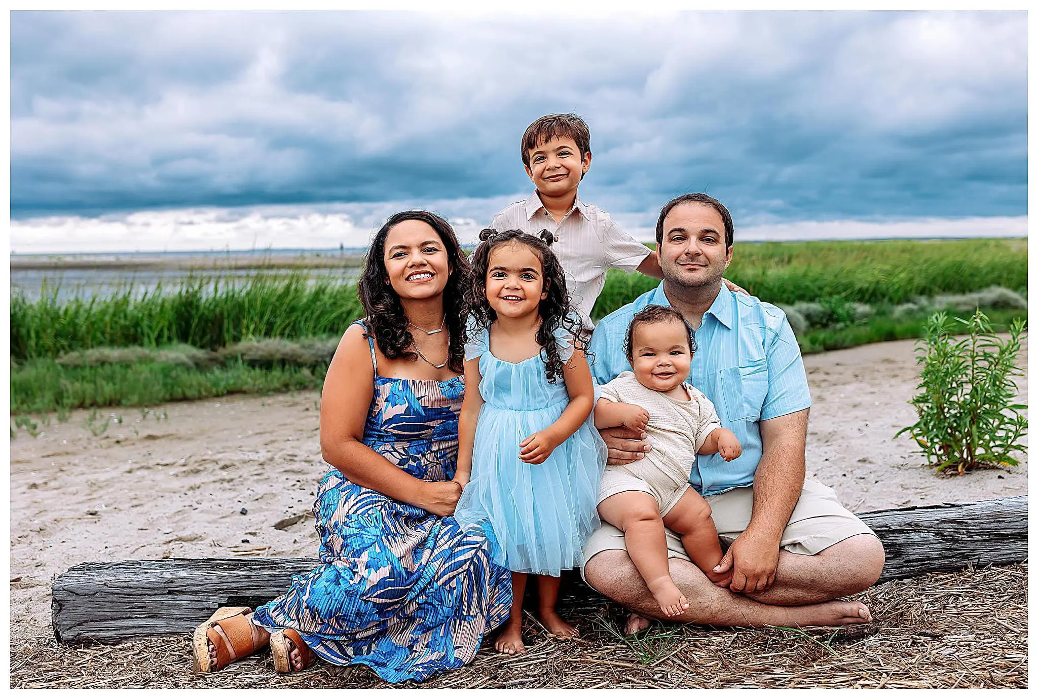 A family of five poses outdoors on a beach. The mother and father sit on a log, with their two daughters in blue dresses between them. A son stands behind, smiling. The background features a grassy shoreline and a cloudy sky.