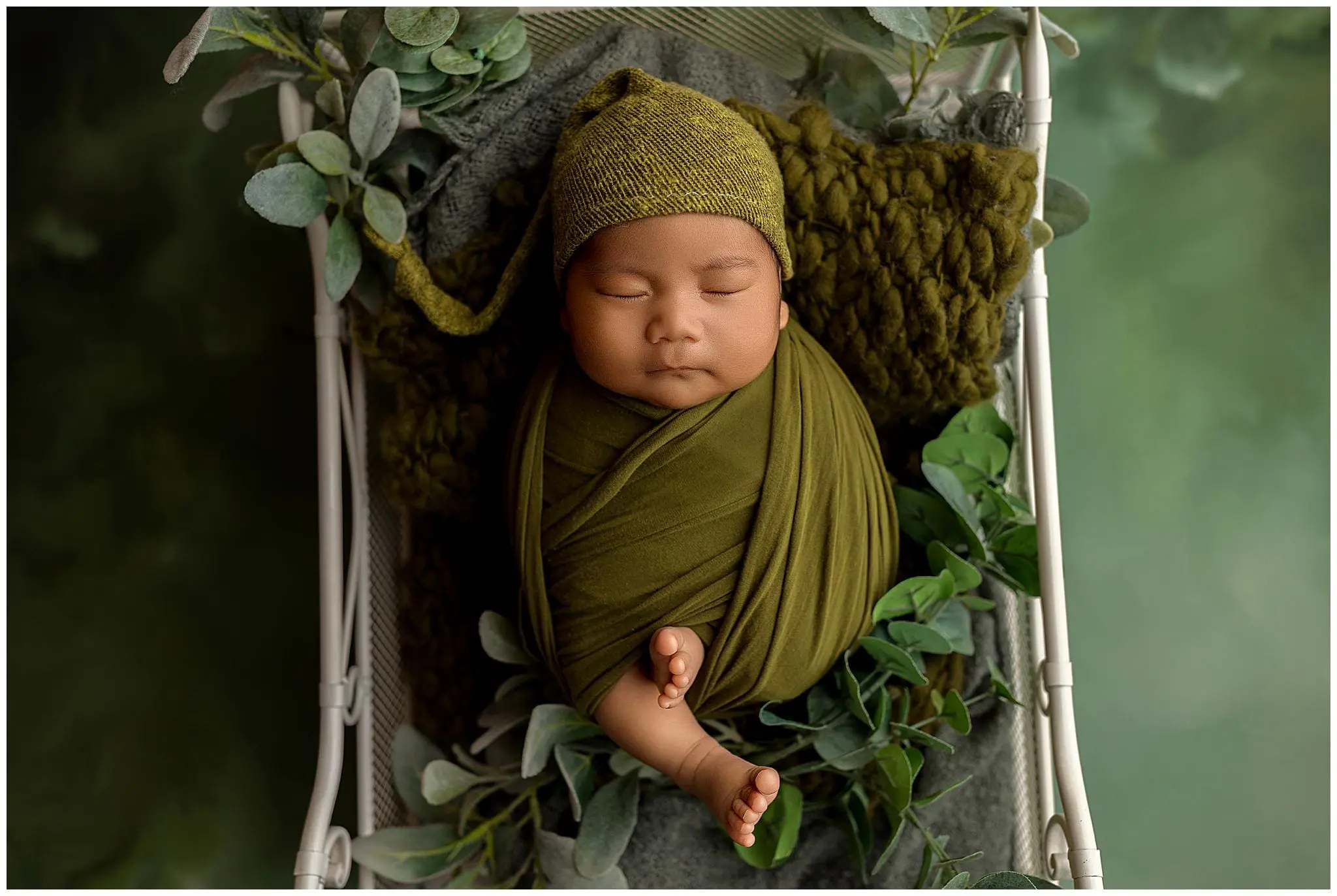 A peacefully sleeping baby is lovingly swaddled in an olive green wrap with a matching knit hat. The serene setting features the baby nestled in a basket amidst lush green foliage, all against a soothing green backdrop.