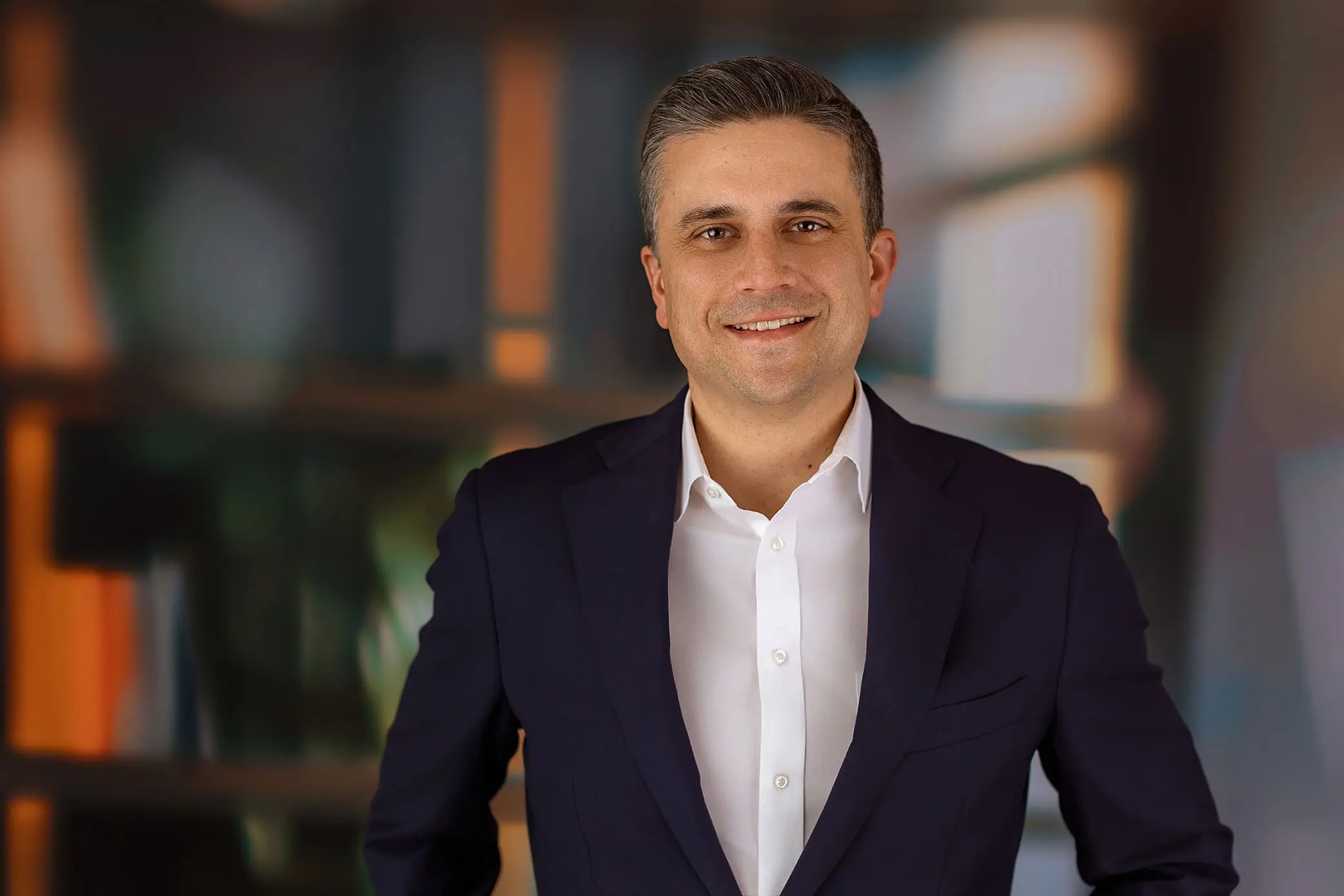 A man in a dark suit and white shirt stands indoors, smiling at the camera. The background is blurred with shades of blue, orange, and green during his Connecticut branding session.
