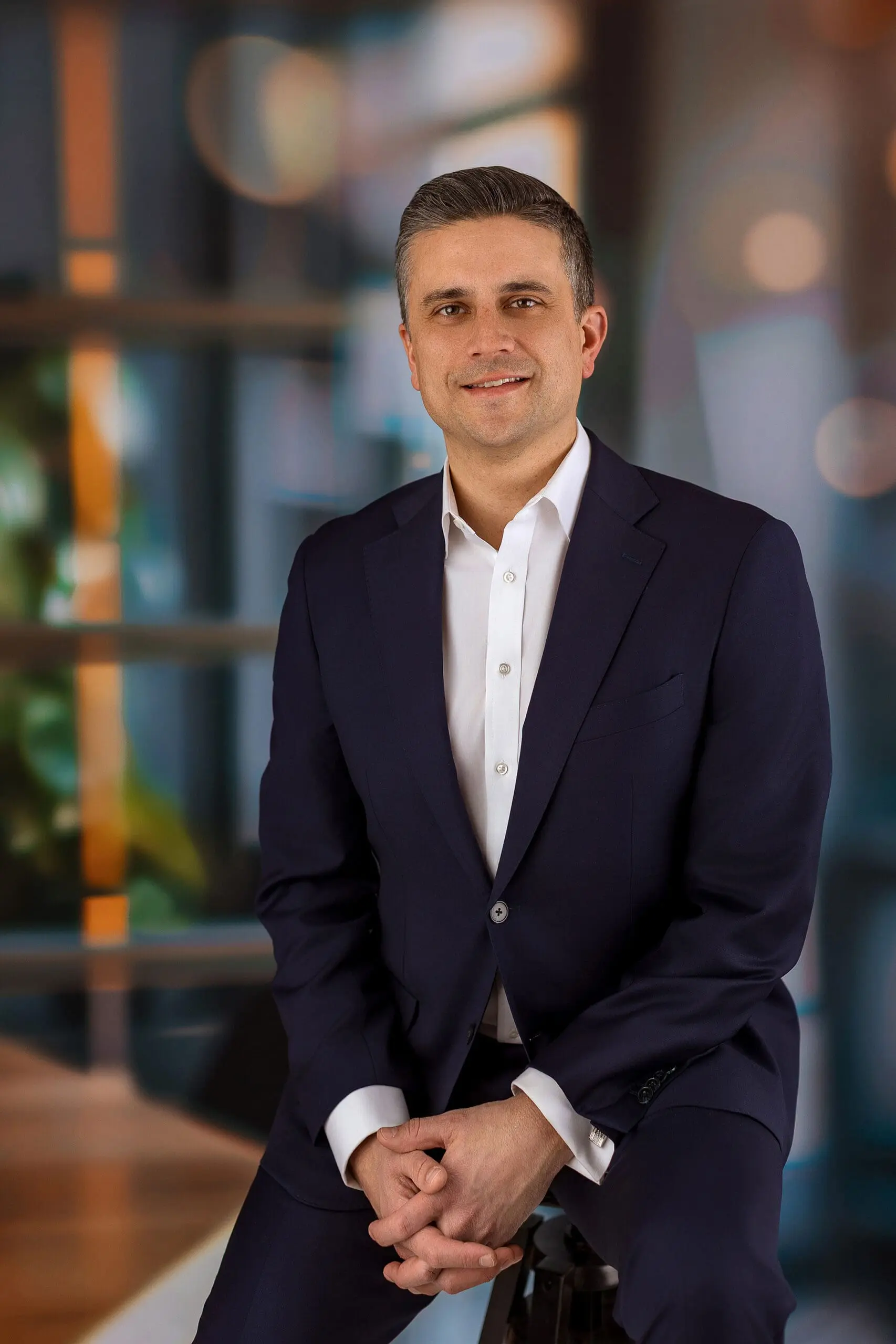 A man in a dark suit and white shirt sits on a stool against a blurred background. The setting appears professional, with soft lighting and bokeh effects. He is smiling gently and has short, gray hair photographed by Connecticut photographer.