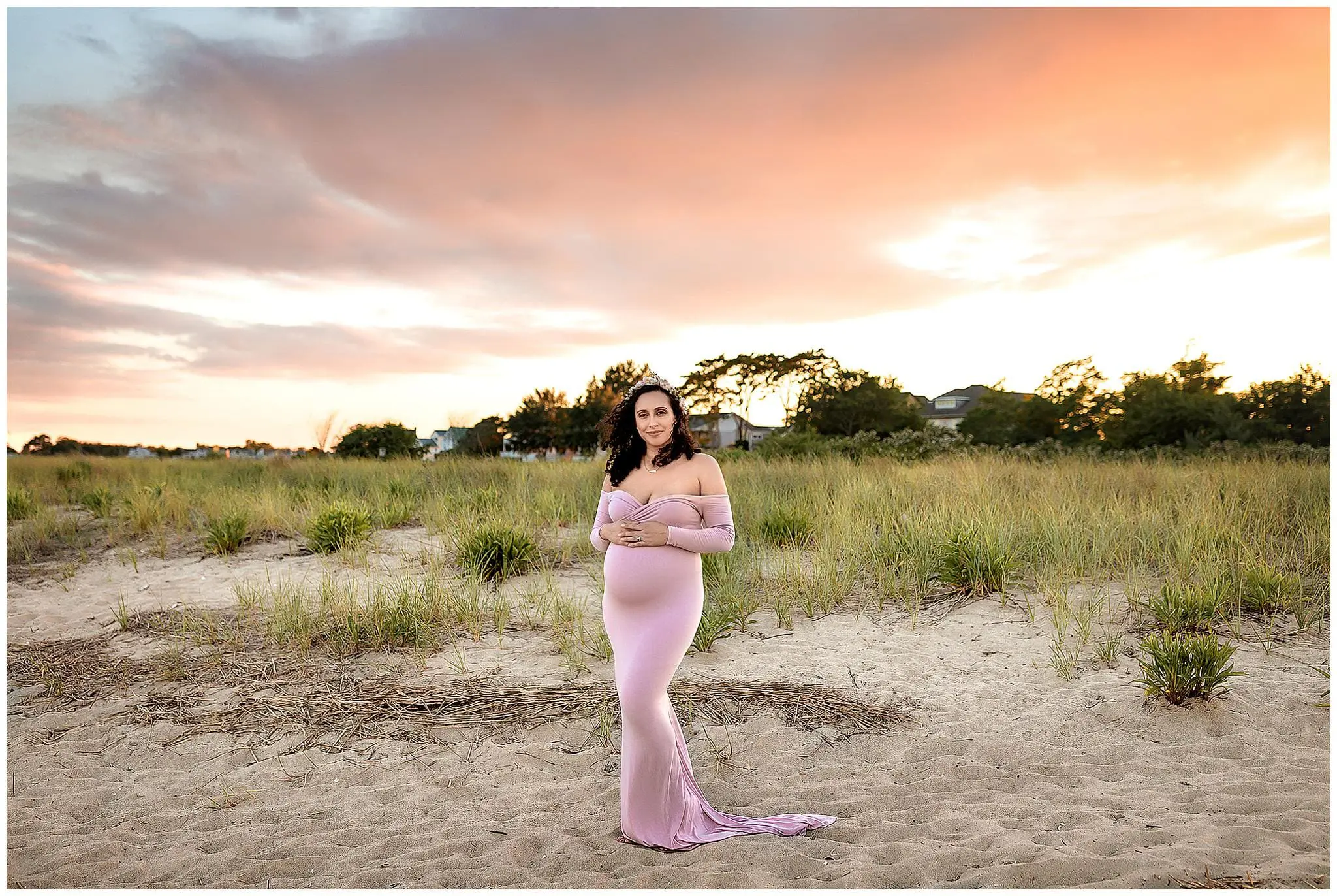 Pregnant woman in a long, flowing pink dress stands on a sandy beach at sunset. The sky is orange and pink with scattered clouds, and greenery is visible in the background. She holds her belly and smiles gently.