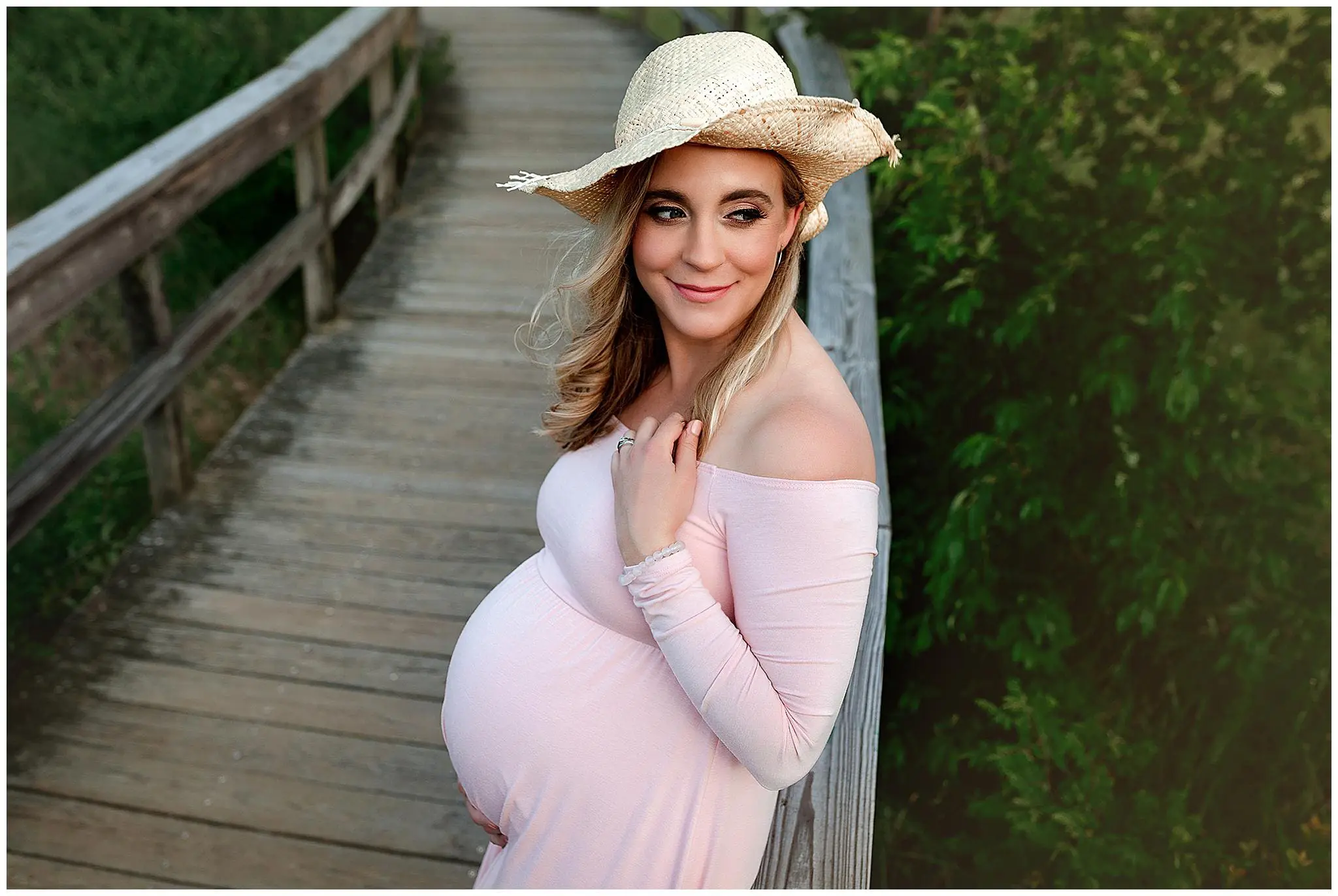 Pregnant woman in a pink dress and straw hat stands on a wooden bridge, smiling with one hand on her belly. She is framed by greenery in the background.