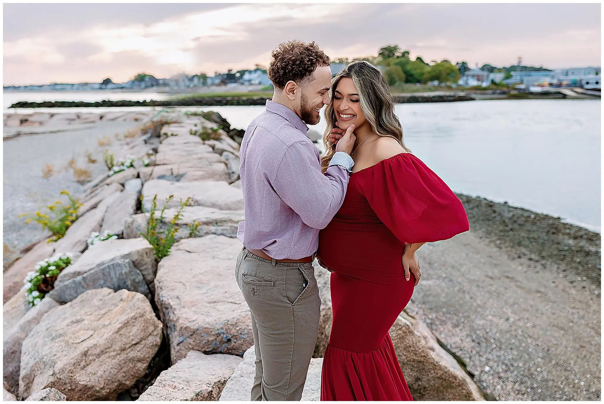 An expecting couple stands on a rocky path by the water in Connecticut, smiling lovingly at each other. The woman, in a flowing red dress, touches the mans face. The sky is a soft pink, and the shoreline is visible in the background.