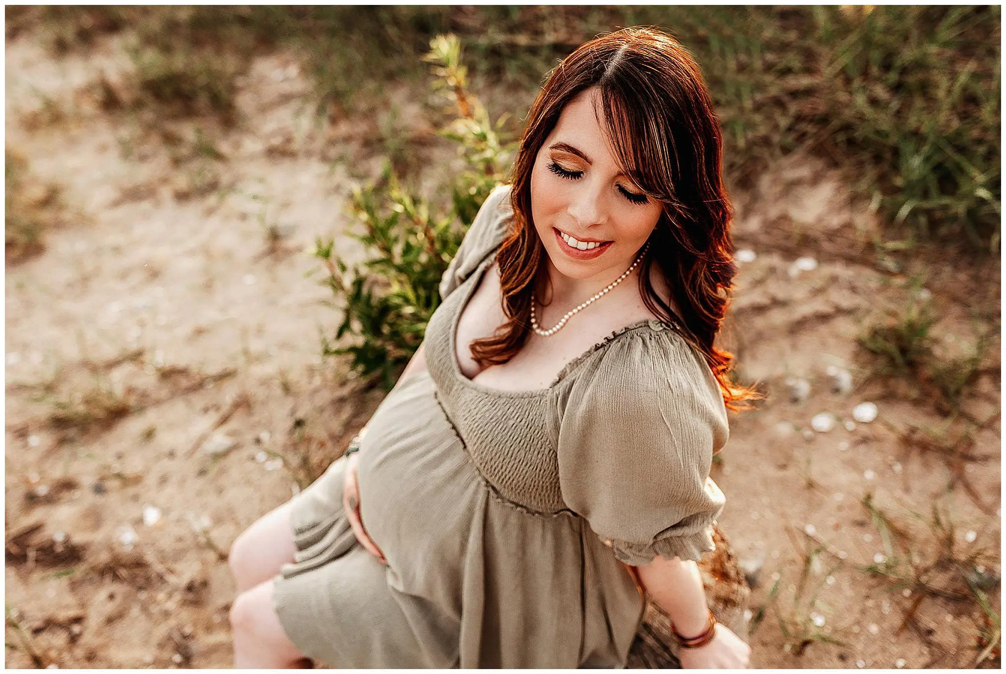 A pregnant woman with long red hair sits outdoors on sandy ground, wearing a light green dress. She smiles softly while looking down. Green plants are nearby, and the warm sunlight enhances her serene expression.