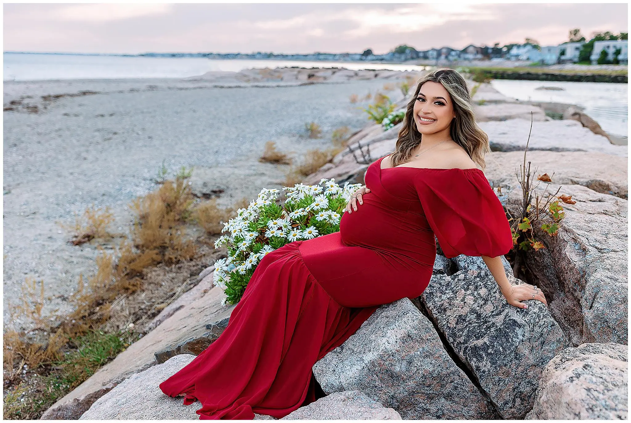 Pregnant woman in a long, off-shoulder red dress sits on rocks by a beach. She is smiling, with one hand on her belly and the other resting on the rocks. White flowers are beside her, and the sea and houses are in the background under a cloudy sky In Connecticut.