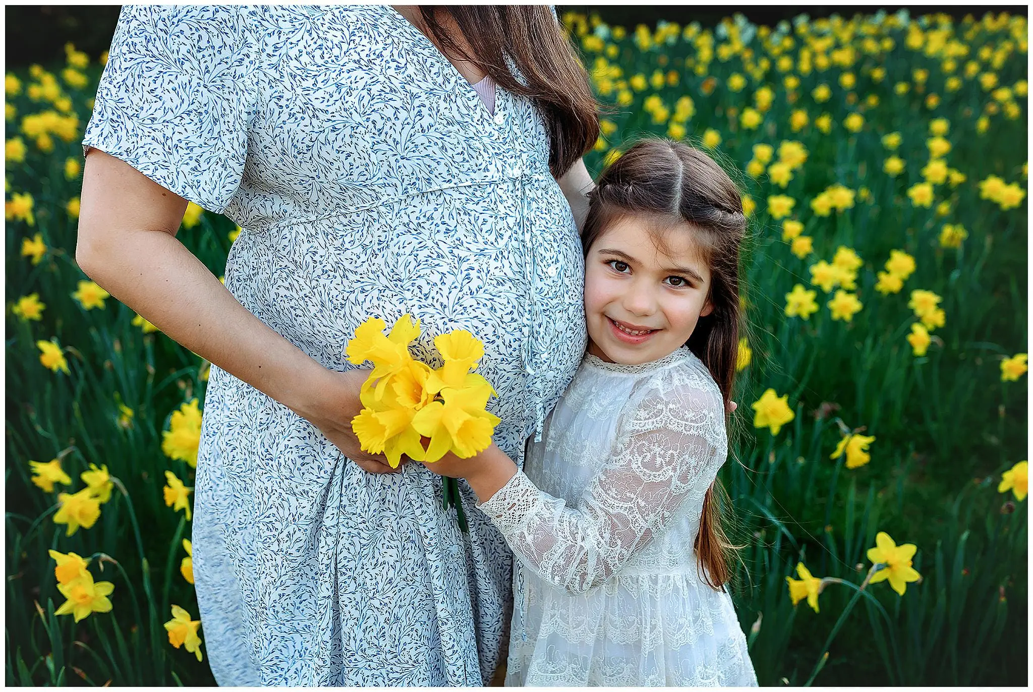 A pregnant woman in a patterned dress stands in a field of yellow flowers, holding a bouquet. A smiling young girl in a white lace dress hugs her and touches the belly. They are surrounded by blooming flowers.