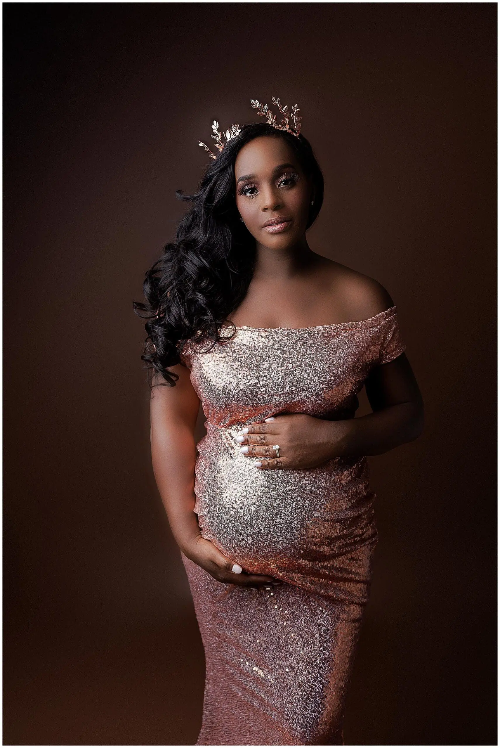 A pregnant woman in a sparkly, off-shoulder rose gold dress poses against a dark background. She wears a matching crown with floral accents and gently cradles her baby bump, looking directly at the camera.