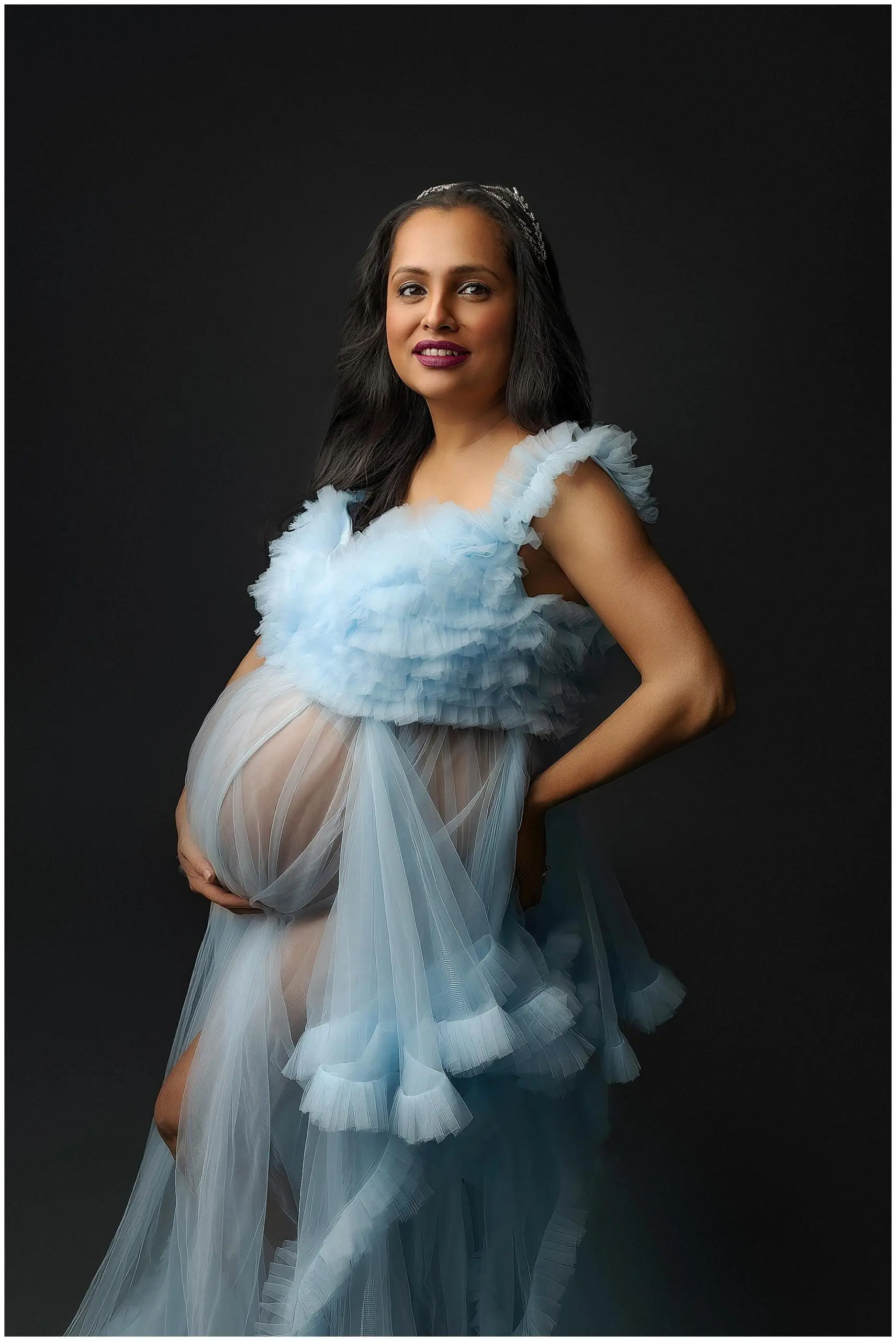 A pregnant woman in a sheer, light blue gown with ruffles poses gracefully against a dark background. Her long, slightly wavy dark hair complements her subtle, confident smile. With hands gently resting on her belly, she embodies the elegance of Connecticut maternity.
