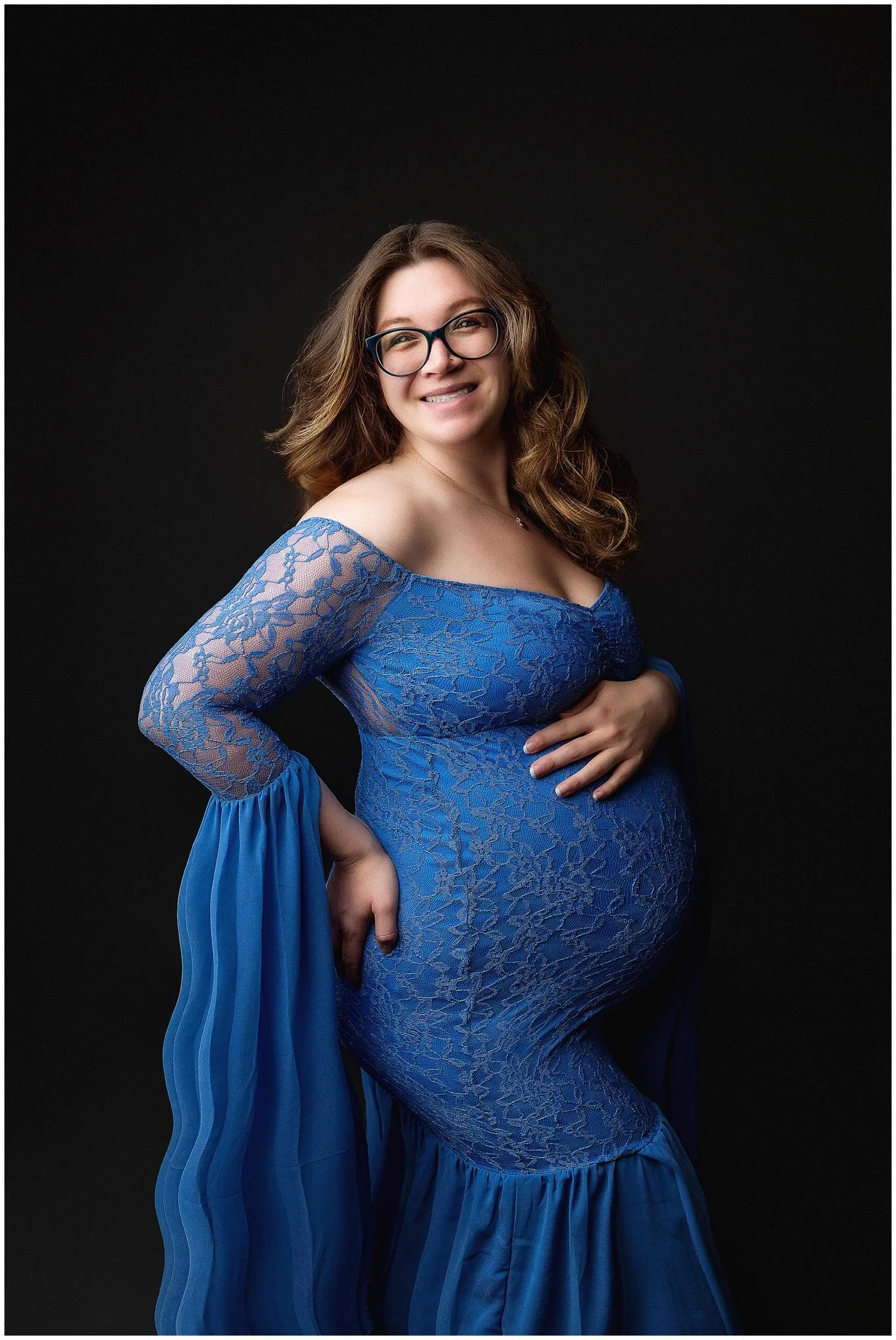 Pregnant woman in a form-fitting blue lace dress poses against a dark background, smiling and resting one hand on her hip and the other on her belly. She wears glasses and has wavy hair cascading over her shoulders during her maternity photo session.