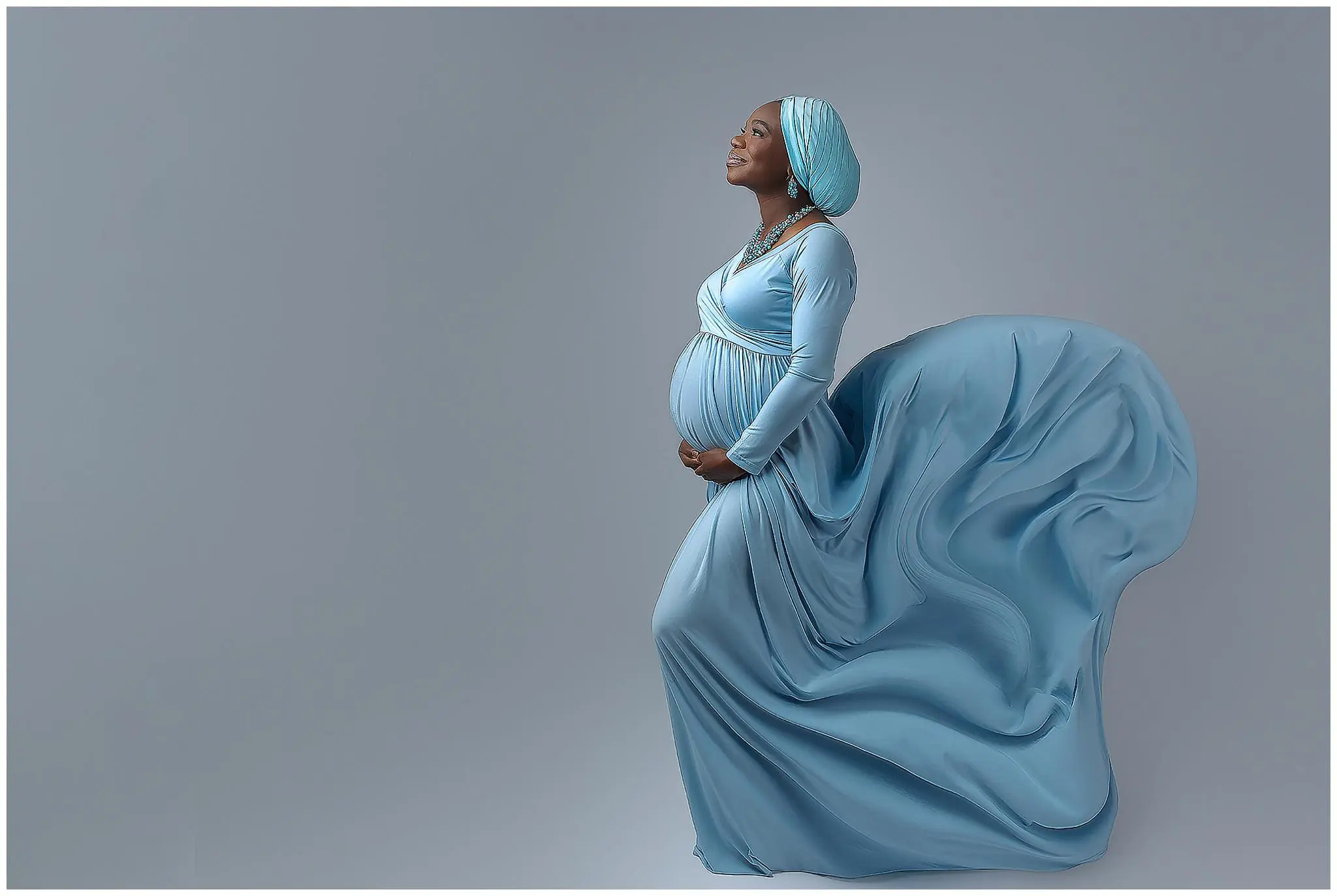 Pregnant woman in a flowing light blue gown and matching headwrap, standing against a gray background. She cradles her baby bump with both hands and gazes upward, exuding grace and serenity. Her gown billows elegantly behind her during her maternity photo session