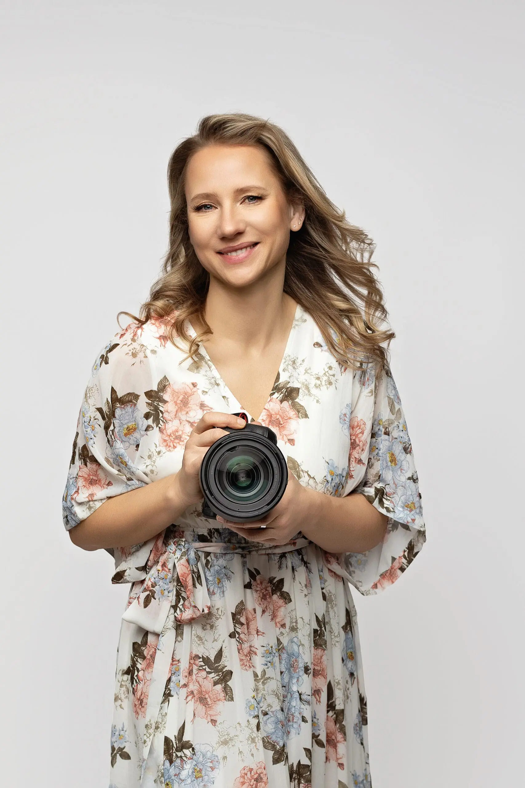 Smiling woman with wavy hair wearing a floral dress holds a camera in front of her against a plain background for her portrait session in Milford.