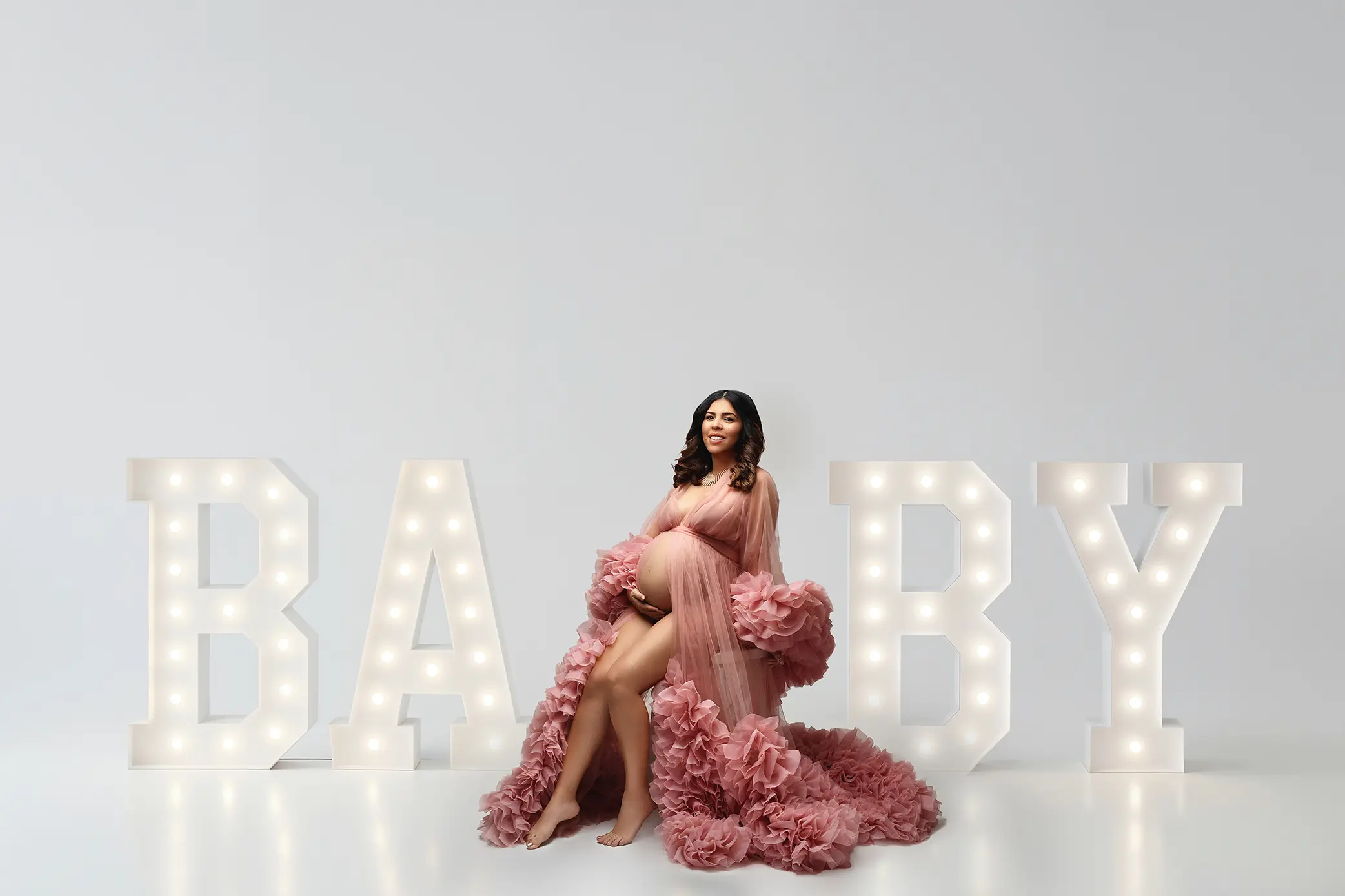 A pregnant woman in a flowing pink gown sits in front of large, illuminated letters spelling BABY against a plain white background.