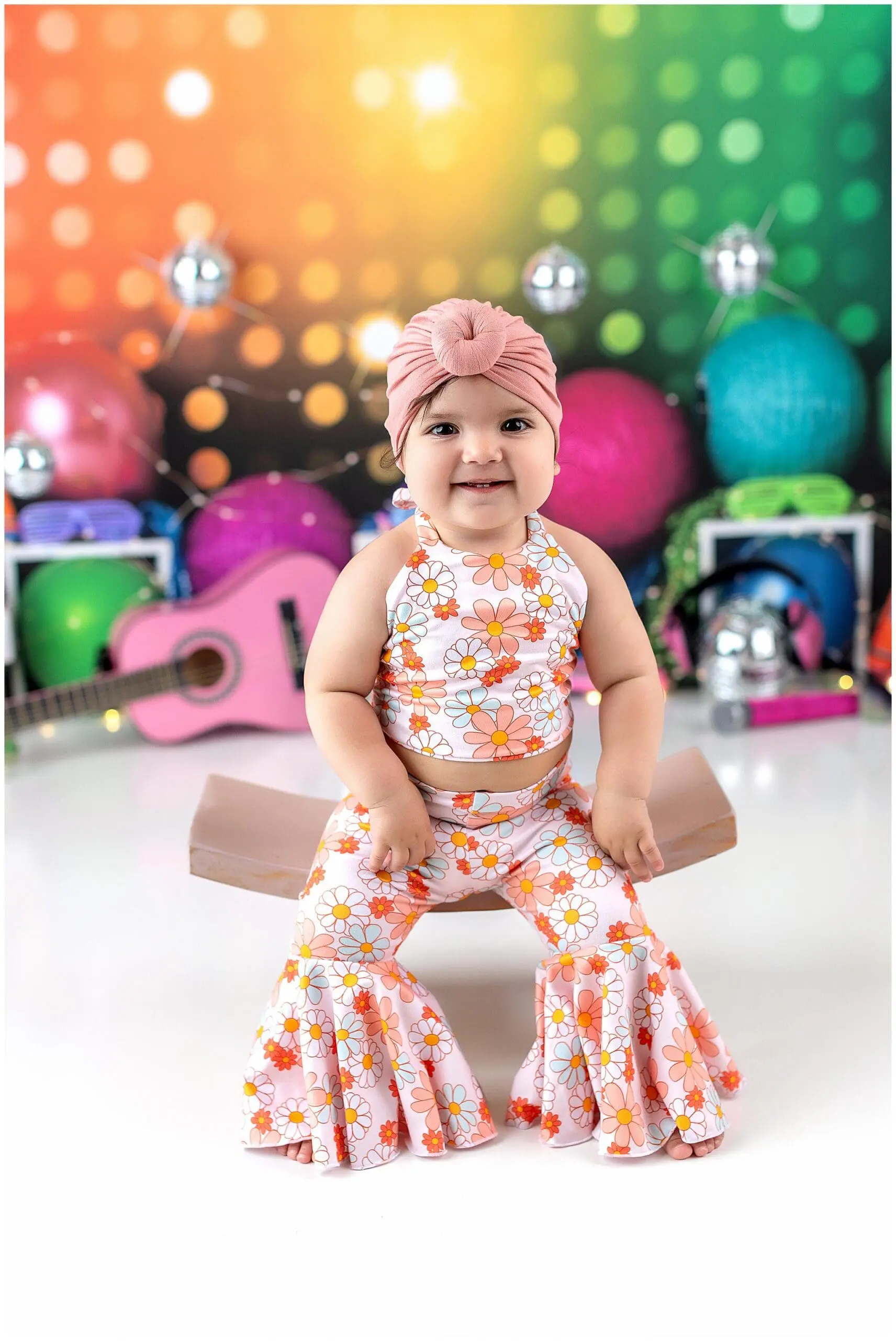 A baby in a floral top and bell-bottom pants sits on a small brown chair, smiling amidst a vibrant cake smash photo setting, complete with a colorful rainbow backdrop, bokeh lights, balloons, a pink guitar, and disco balls.