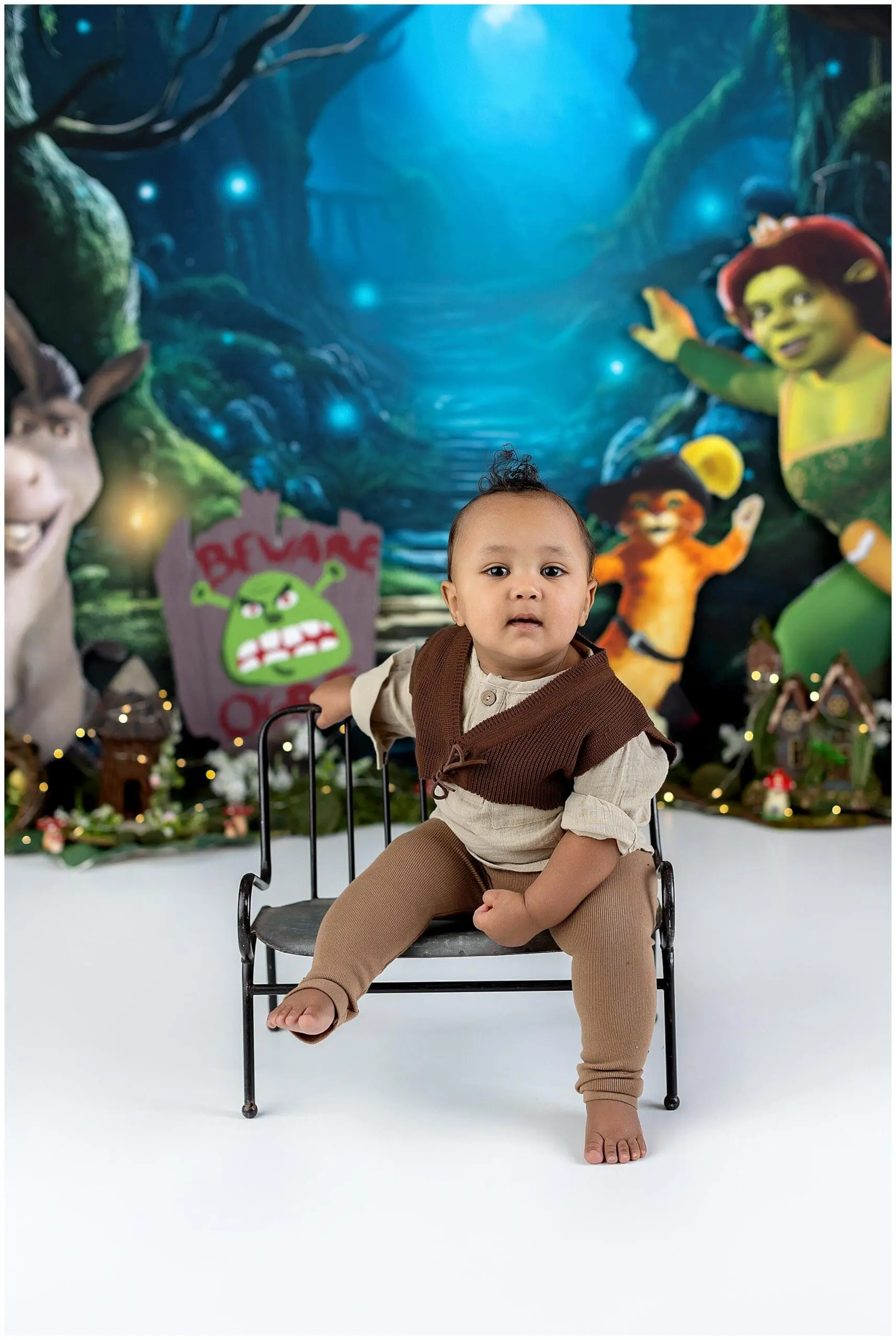 A baby sits on a small bench wearing a brown vest and leggings. The background is a colorful, fantasy-themed scene with cartoon characters and whimsical decorations, including a Beware sign with a green face.