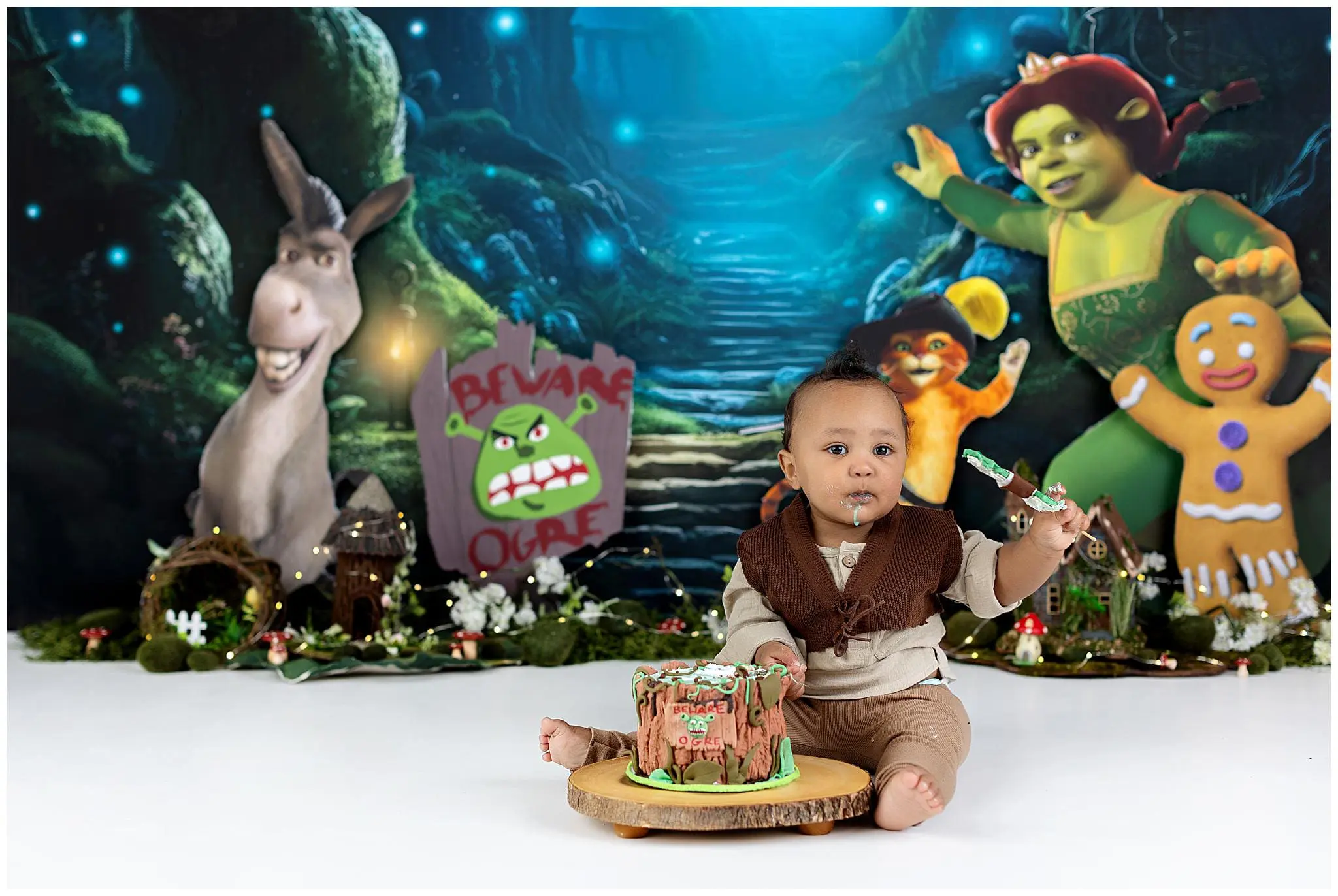 A baby dressed in medieval-style clothing sits gleefully amidst a Shrek themed cake smash. The partially eaten cake matches the whimsical scene, complete with characters like Donkey and Shrek cheerfully gracing the backdrop.