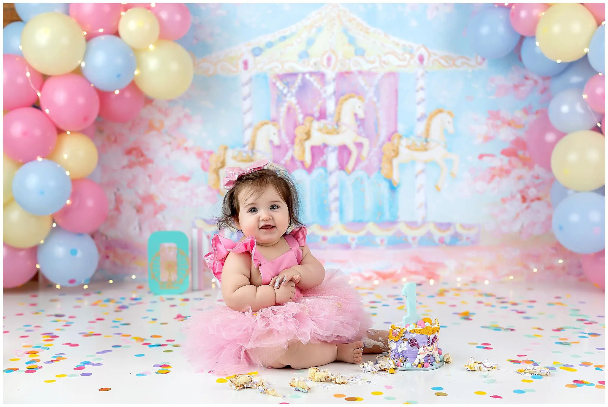 A baby in a pink tutu sits on a colorful confetti-covered floor, surrounded by balloons. A carousel backdrop adds whimsy, while a small cake with a candle hints at an upcoming cake smash to celebrate the birthday.