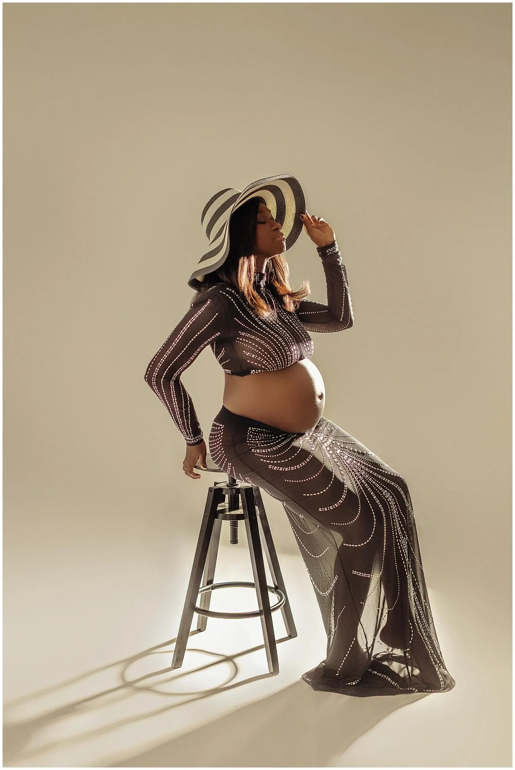 Pregnant woman in a sheer, patterned dress and striped hat sits on a stool, side profile visible, against a neutral background. Her hand gently holds the brim of her hat while posing for her maternity session.