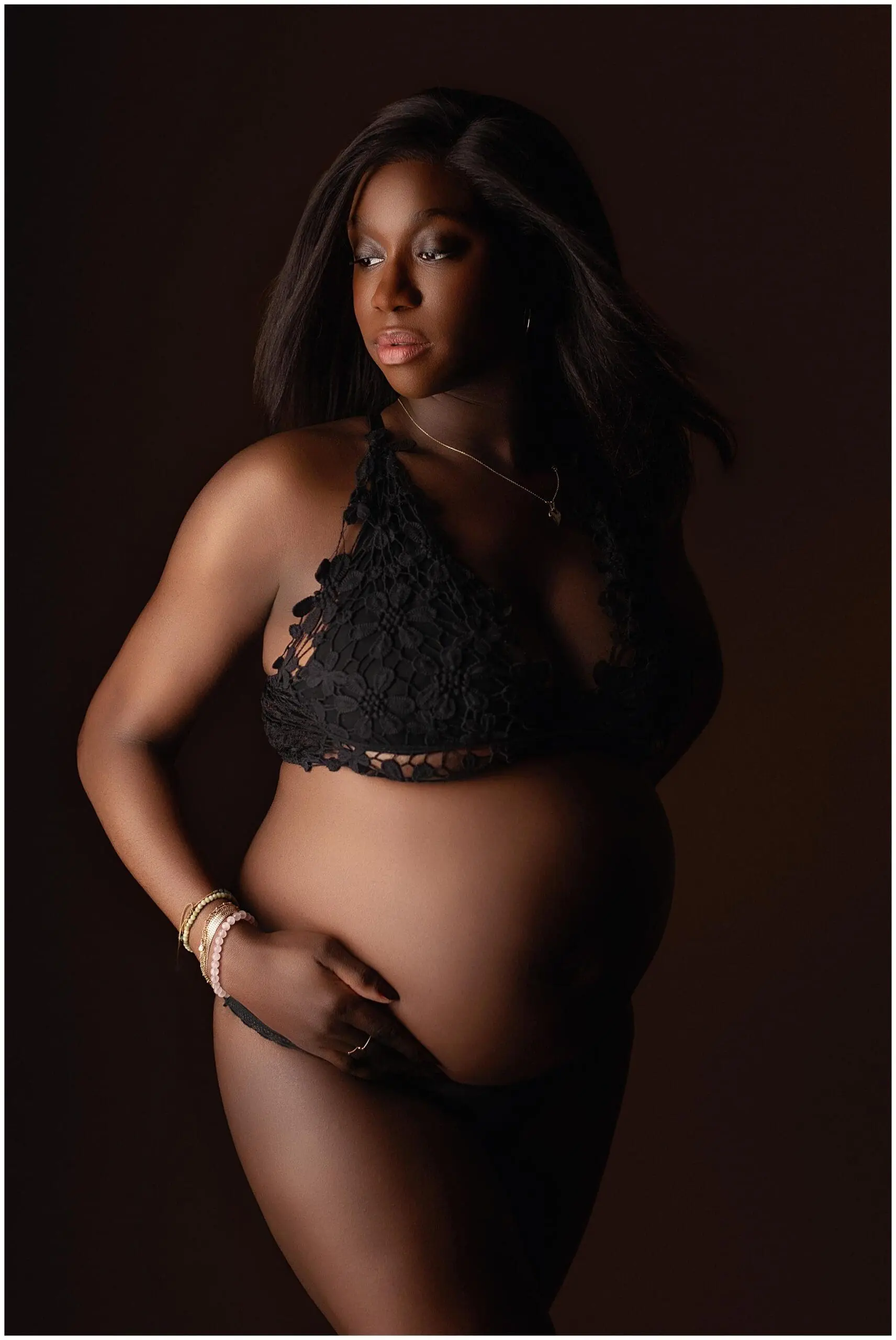 Pregnant woman in black lace lingerie stands confidently against a dark background. She gazes thoughtfully to the side, her long hair cascading over her shoulders. Her posture highlights her baby bump, with one hand on her side.