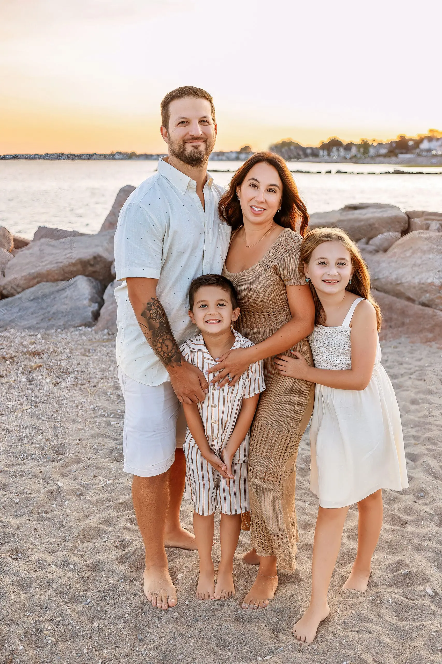 A family of four stands on a sandy beach with large rocks in the background. The man wears a light shirt and shorts, the woman a brown dress, and the two children are in striped and white dresses. They smile at the camera during sunset at their family photographer in Connecticut.
