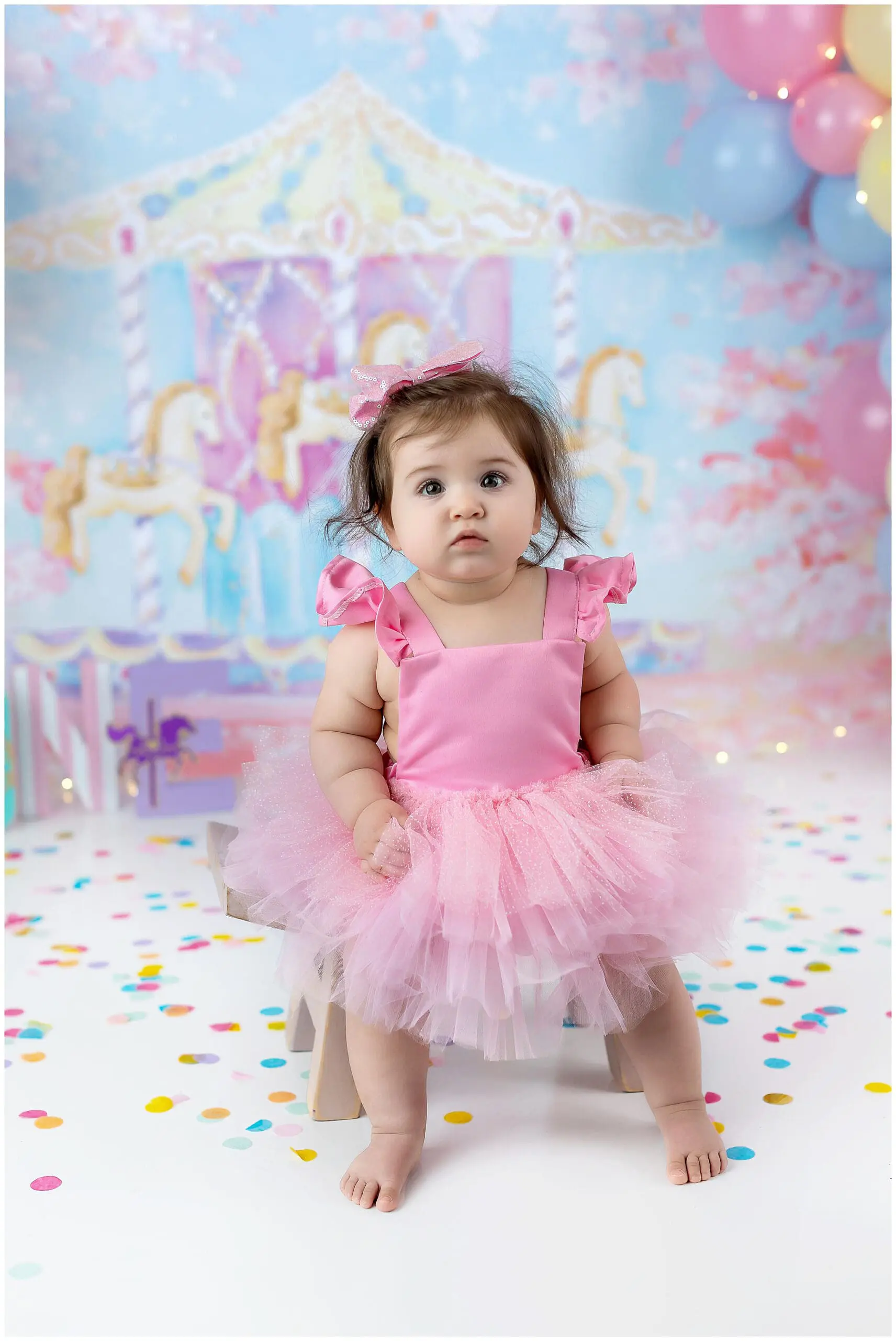 A toddler wearing a pink dress and tulle skirt sits on a small wooden bench. The background features a pastel carousel with balloons and confetti scattered on the floor. The child has a curious expression during her Ct cake smash session.