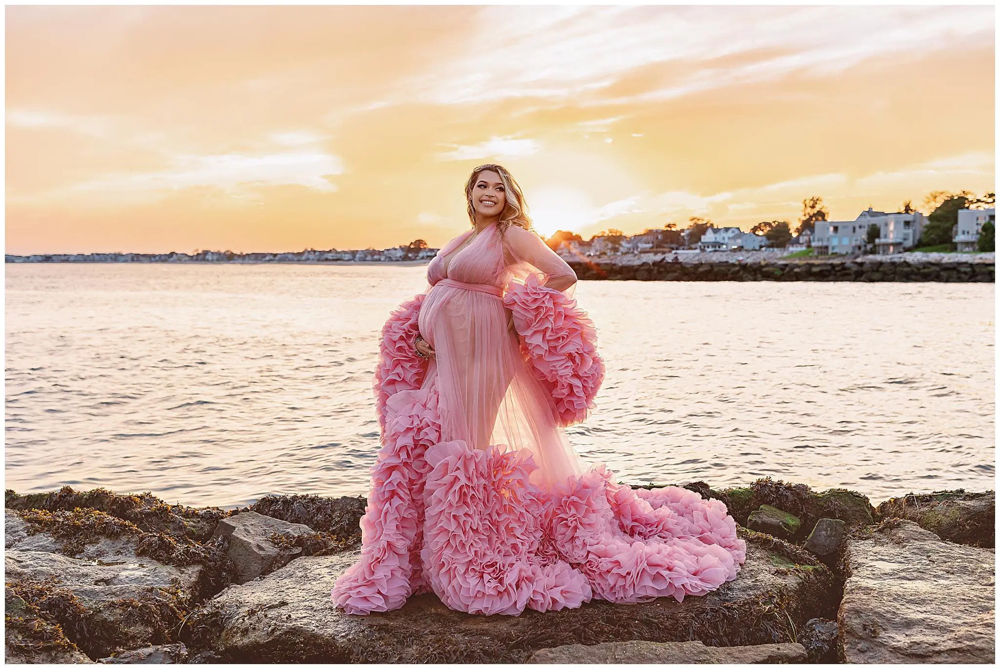 A person in a flowing pink dress stands on rocks by the water at sunset. The sky is a blend of orange and pink hues, reflecting on the calm water. Houses are visible along the shoreline.