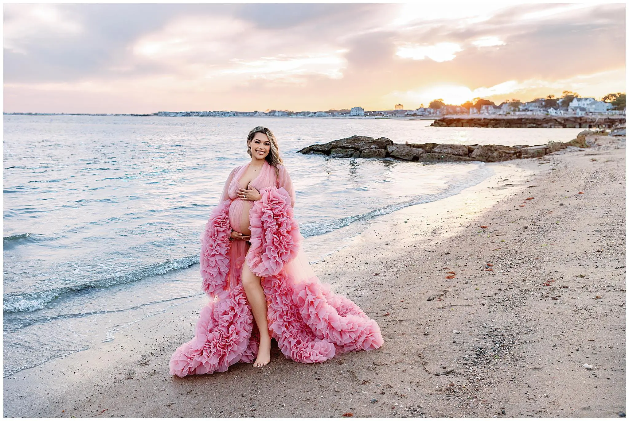 A woman stands on a sandy beach at sunset, wearing a flowing pink gown. She is barefoot with waves gently lapping at the shore. The sky is partly cloudy with orange and pink hues, and there are houses visible in the distance.