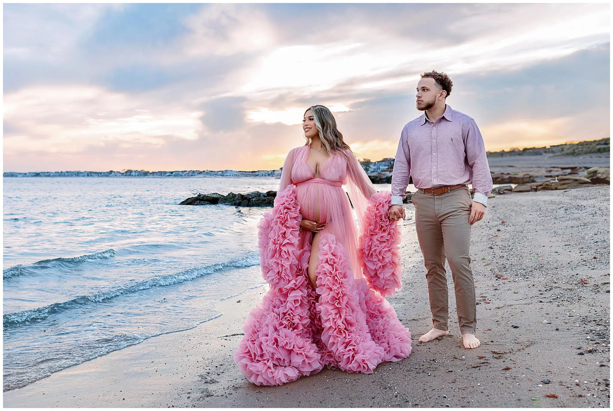 A maternity couple stands barefoot on a beach, the woman in a flowing pink gown and the man in a matching pink shirt with beige pants. They hold hands, gazing towards the ocean as the sunset paints the sky in stunning hues.