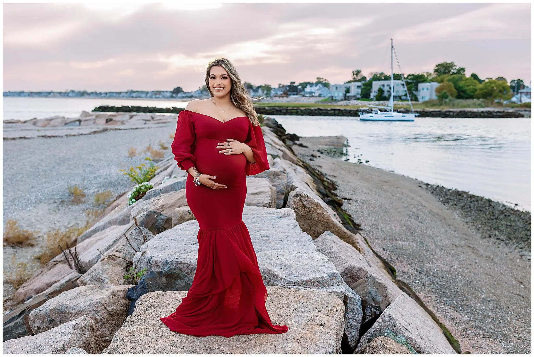 A pregnant woman in a flowing red dress stands on large rocks by the water, smiling and holding her belly. In the background, a calm Connecticut sea frames a small sailboat and houses along the shoreline. The sky is slightly overcast, evoking a serene coastal charm during her ct maternity photo session.
