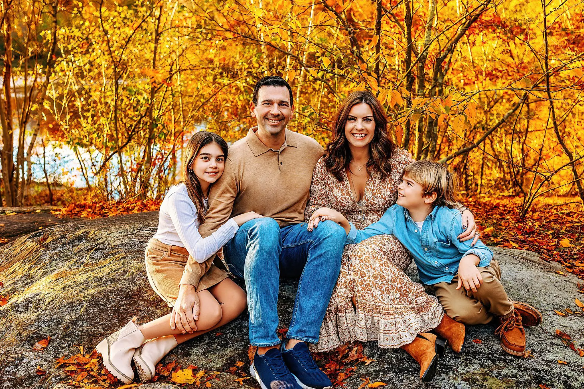 A family of four poses on a large rock amidst vibrant autumn foliage in Connecticut. The parents are in the center, with their daughter on the left and son on the right. Smiling in earthy tones and surrounded by bright orange leaves, it’s a perfect snapshot of family photos in Connecticut.