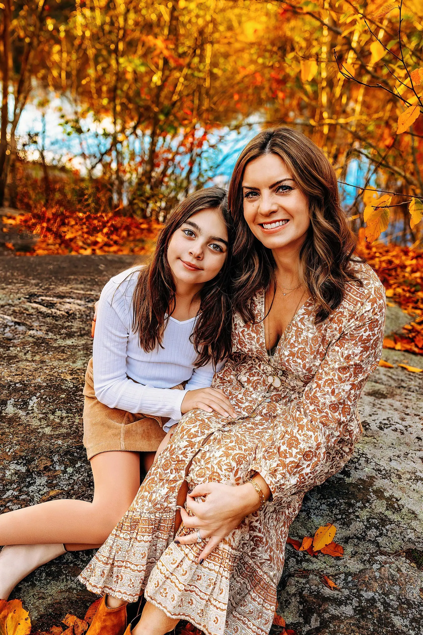 A mom and a young girl sit on a rock in an autumn setting. The background is filled with vibrant fall foliage in shades of orange and yellow. Both are smiling, and the woman wears a patterned dress while the girl wears a white top and beige skirt.